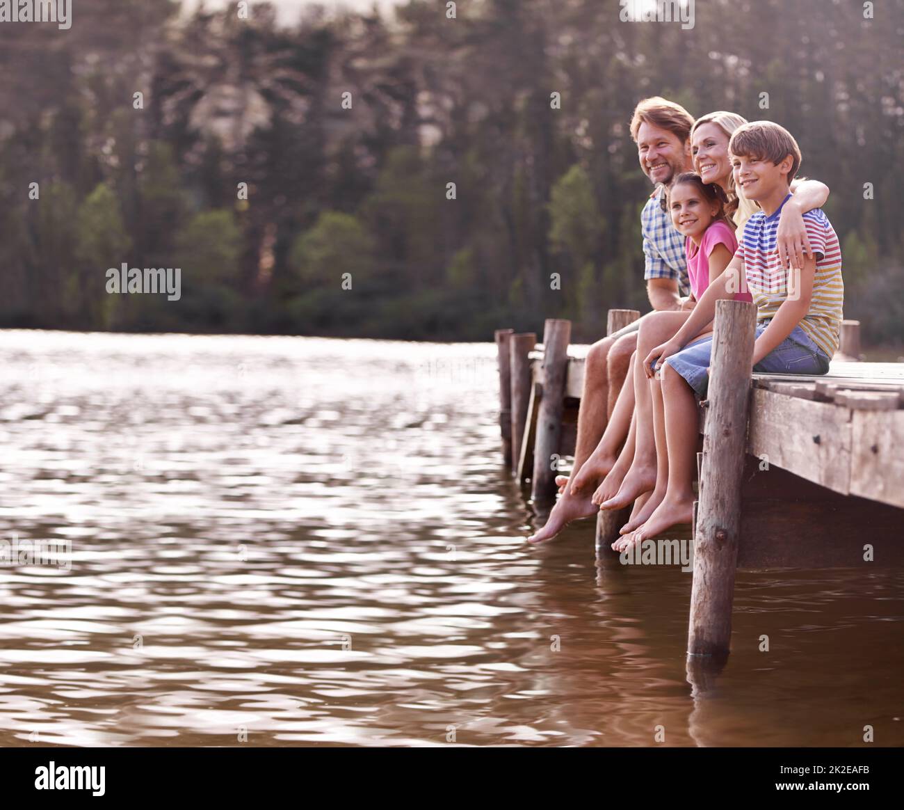 Nichts ist besser als der jährliche Familienurlaub am See. Eine glückliche vierköpfige Familie sitzt auf einem Steg am See. Stockfoto