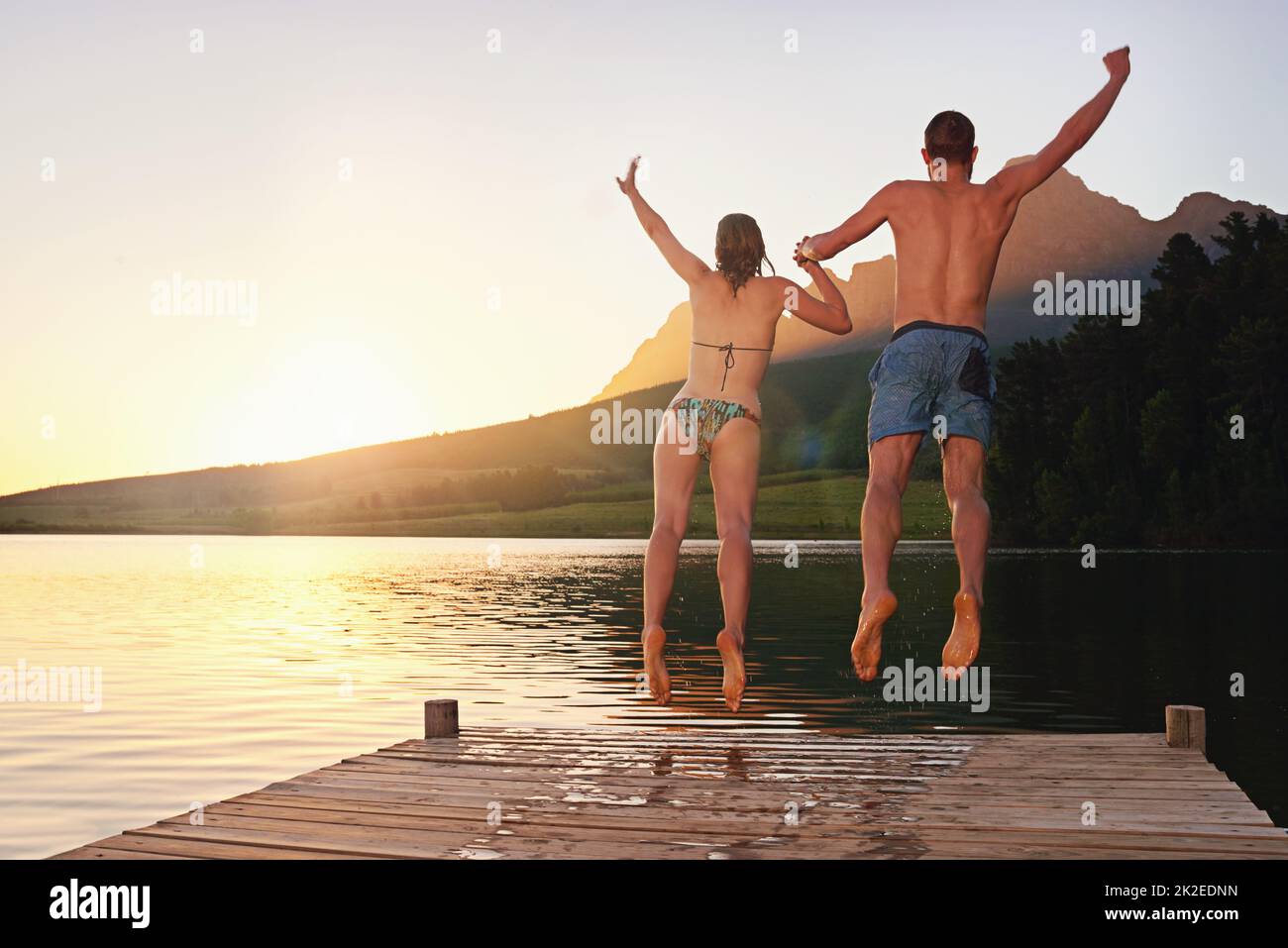Auf dem Sprung. Rückansicht eines jungen Paares in Badeanzügen, das bei Sonnenuntergang von einem Dock springt. Stockfoto
