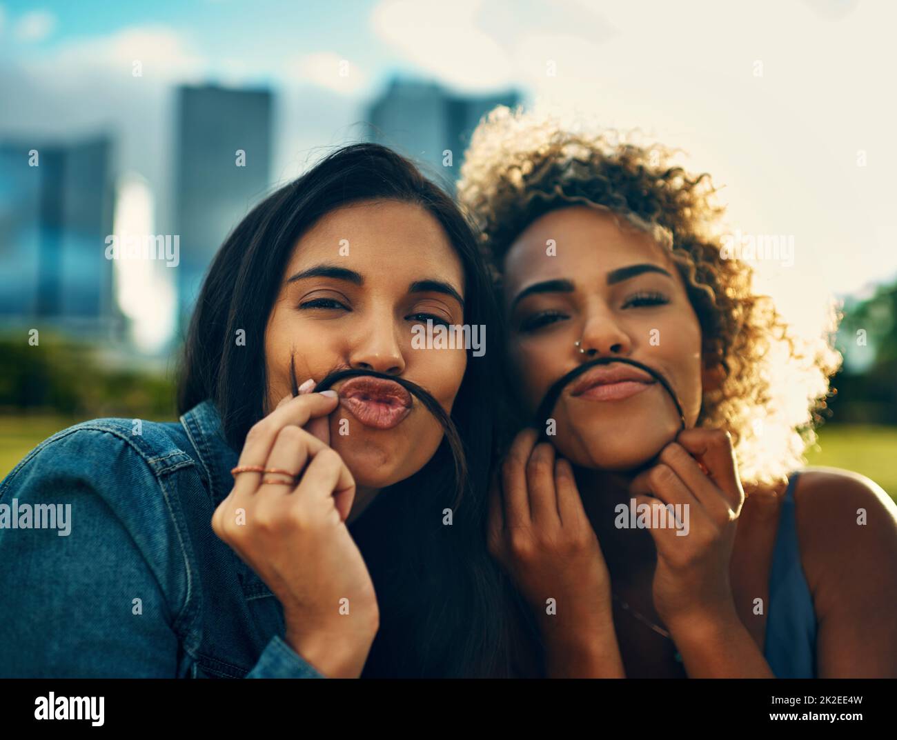 Schließlich fand ich jemanden, der so verrückt war wie ich. Beschnittenes Porträt zweier attraktiver junger Freundinnen, die in einem Park mit ihren Haaren einen Schnurrbart machen. Stockfoto