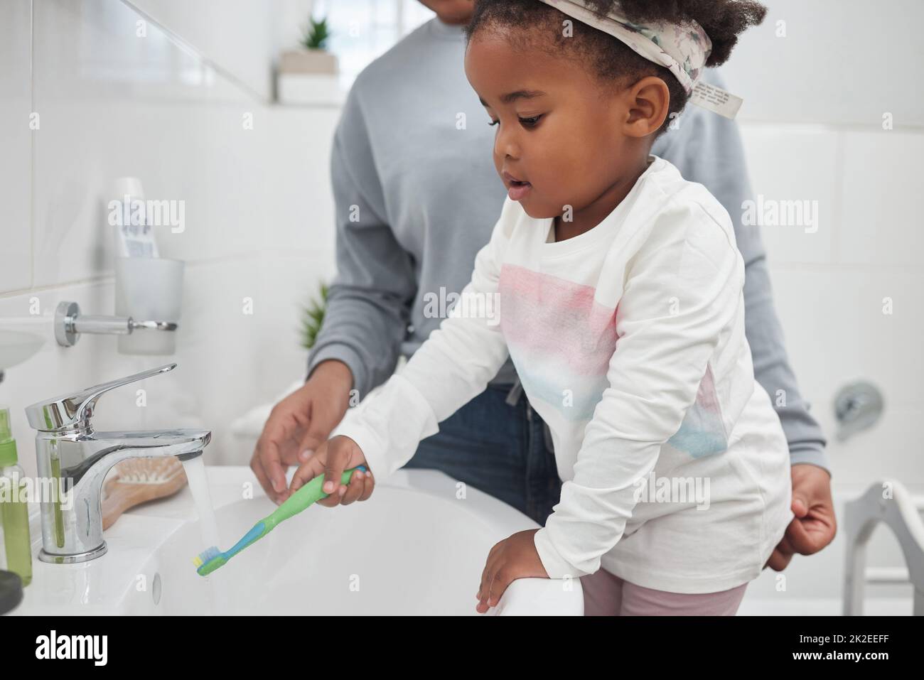 Schon in jungen Jahren alles über gute Hygiene lernen. Aufnahme einer Mutter, die ihrer kleinen Tochter hilft, ihre Zahnbürste zu Hause am Wasserhahn im Badezimmer zu spülen. Stockfoto