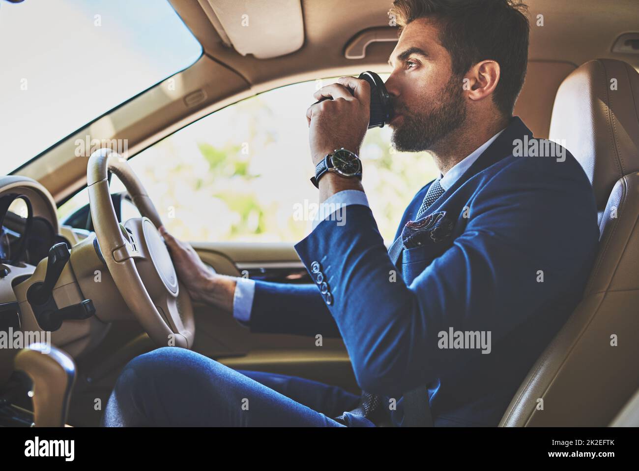 Kaffee hält ihn in Gang. Aufnahme eines fröhlichen jungen Geschäftsmannes, der in seinem Auto zur Arbeit fährt, während er im Verkehr sitzt und Kaffee trinkt. Stockfoto