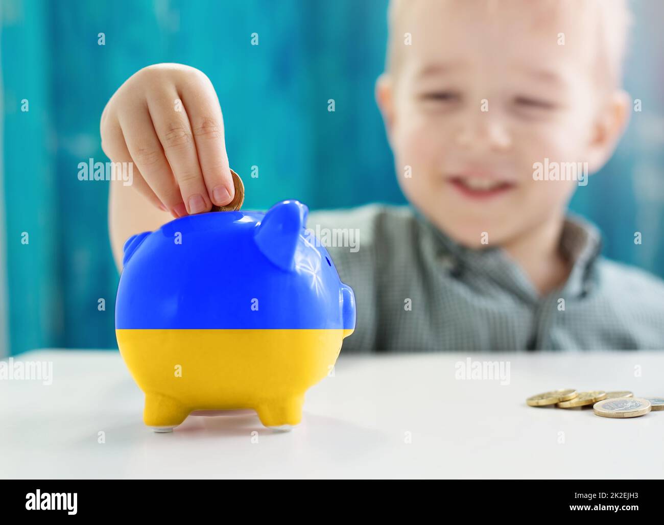 Kind sitzt am Tisch mit Geldmünzen und Biggybank in der Farbe der ukrainischen Flagge. Stockfoto