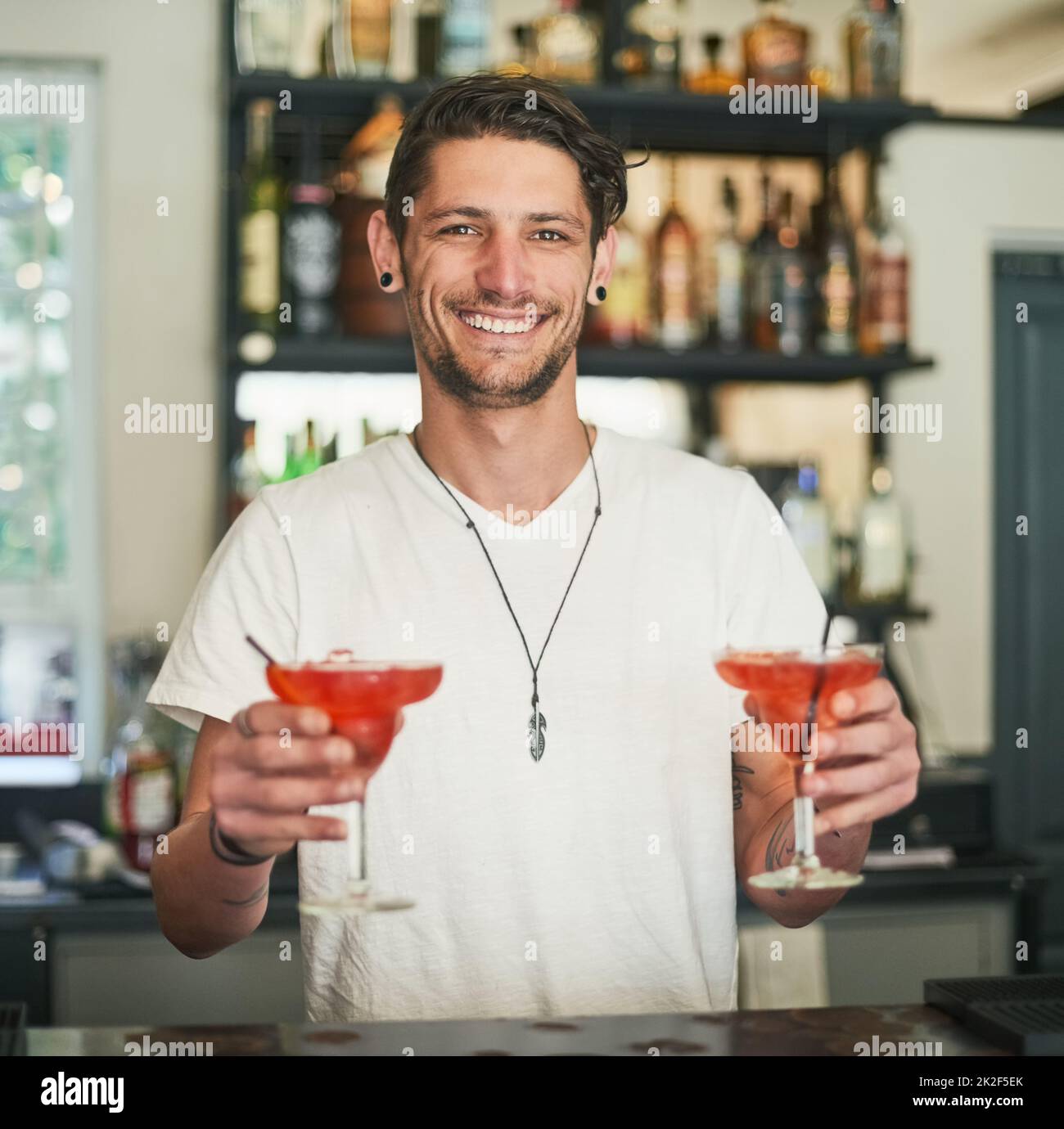 Gemischt nur für Sie. Porträt eines glücklichen jungen Barkeepers, der zwei Cocktails hält, während er hinter der Bar steht. Stockfoto