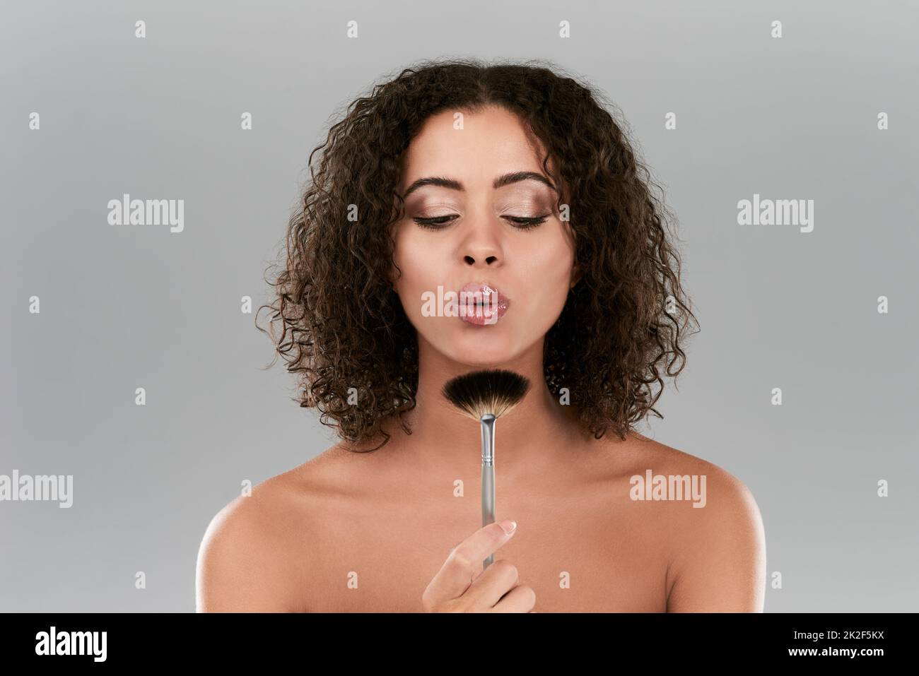 Vor dem Auftragen immer ein wenig abblasen. Studio-Aufnahme einer schönen jungen Frau, die einen Make-up-Pinsel vor einem grauen Hintergrund bläst. Stockfoto