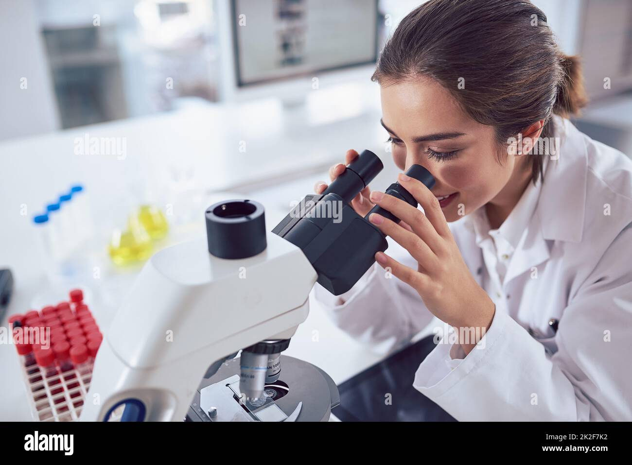 Ich sehe keine Unterschiede in diesem Experiment. Aufnahme einer fröhlichen jungen Wissenschaftlerin, die durch die Linse eines Mikroskops schaut, während sie in einem Labor sitzt. Stockfoto