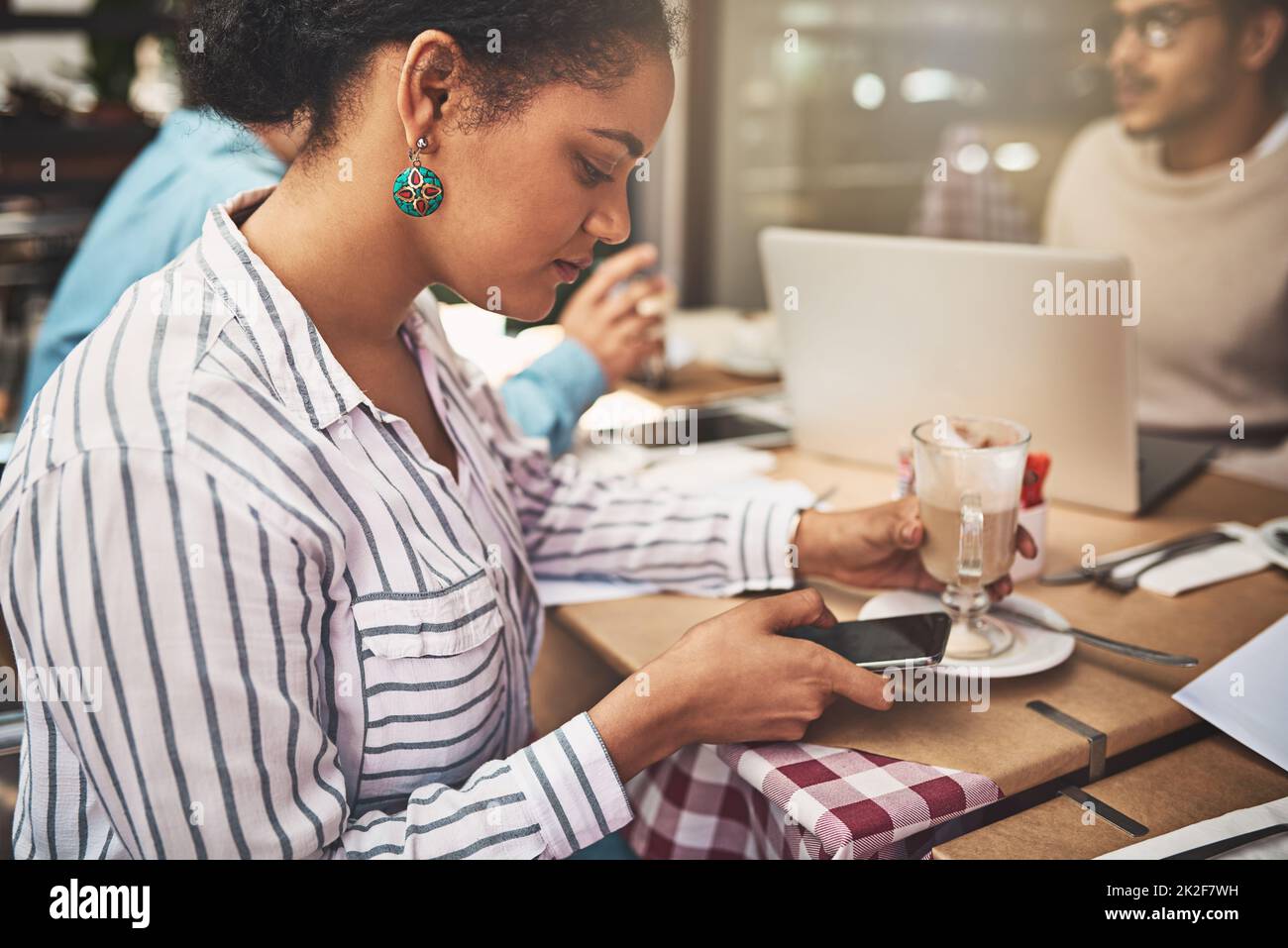 Immer beschäftigt wie üblich. Aufnahme einer fokussierten jungen Frau, die auf ihr Handy schreibt, während sie an einem Coffeeshop sitzt und ein heißes Getränk trinkt. Stockfoto