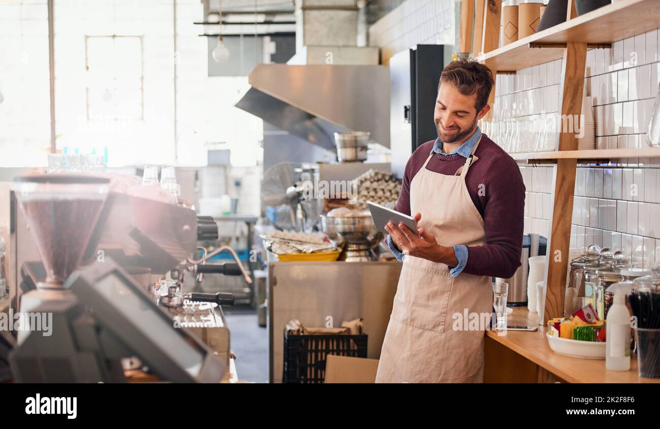 Online-Vergleich von Kaffeemarken. Aufnahme eines glücklichen jungen Geschäftsbesitzers, der ein Tablet benutzte, während er in seinem Café stand. Stockfoto