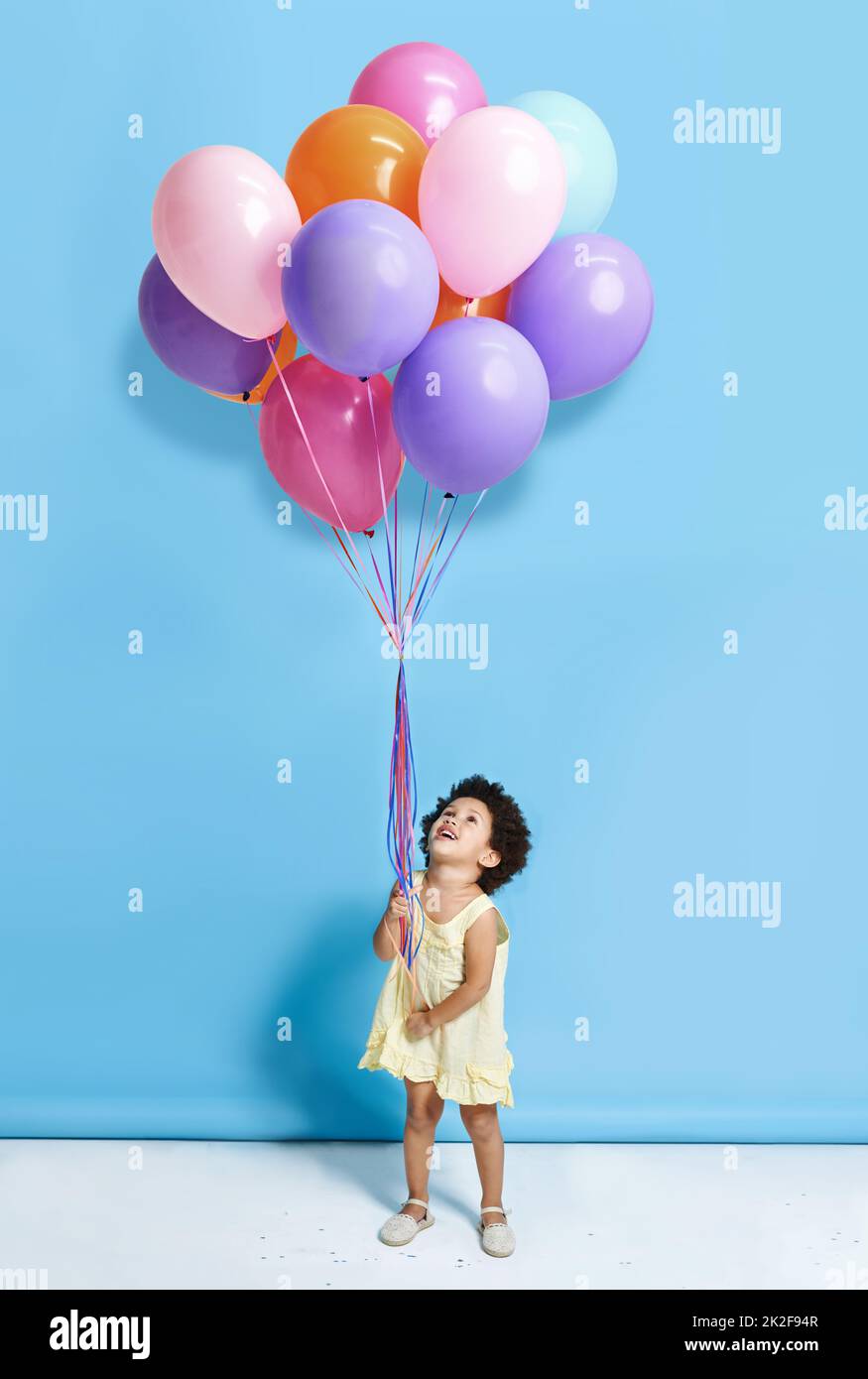 Sollen wir Mr. Balloon tanzen? Aufnahme eines niedlichen kleinen Mädchens mit einem Haufen Ballons vor blauem Hintergrund. Stockfoto