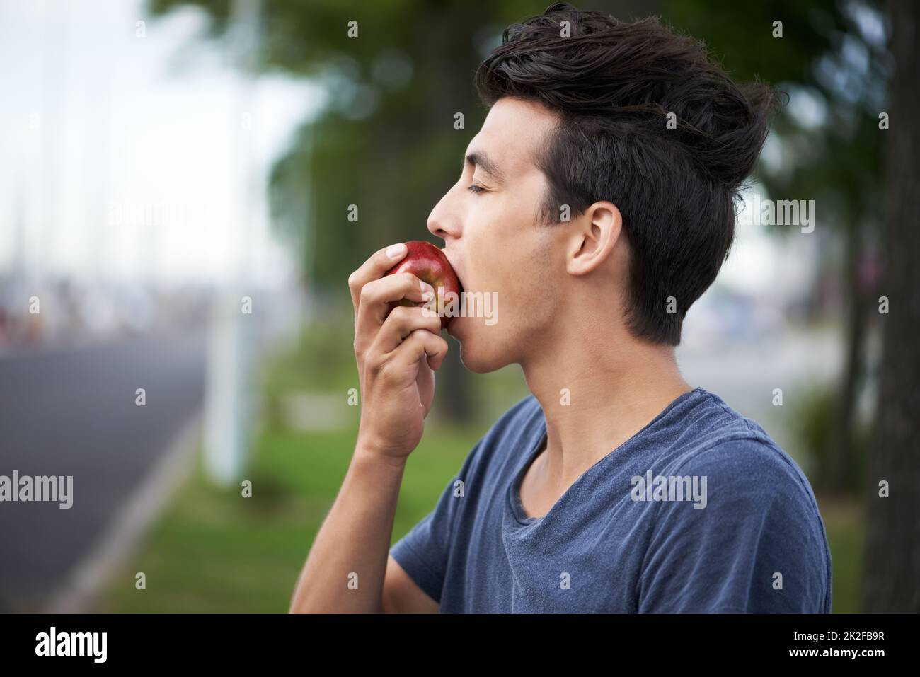 Einen Bissen nehmen. Ein junger Mann, der einen Apfel beißt, während er auf den Bus wartet. Stockfoto