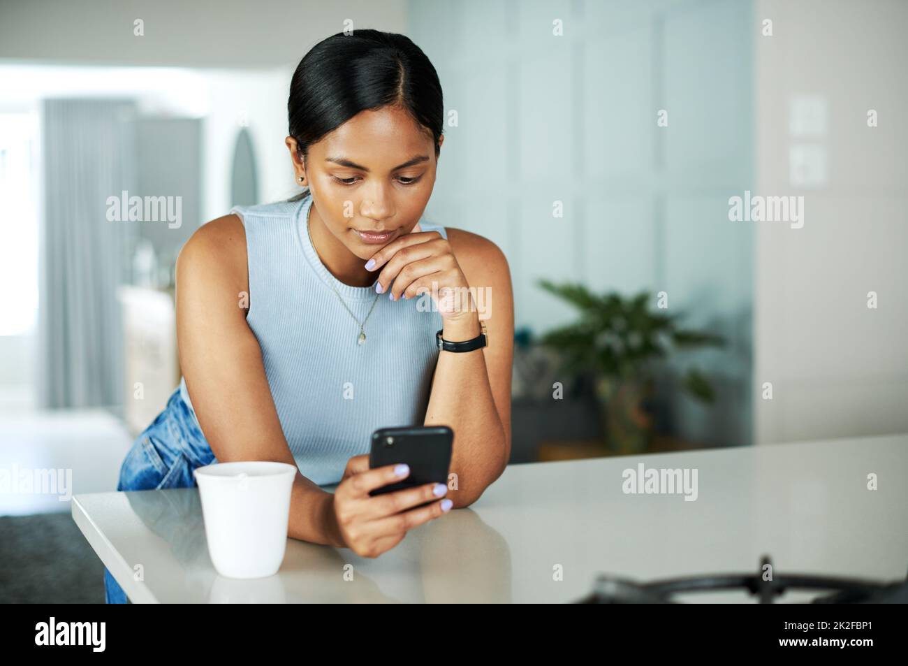 Hmm, das sieht genau richtig aus. Foto einer attraktiven jungen Frau, die sich auf ihren Küchentisch lehnt und bei der Benutzung ihres Handys nachdenklich aussieht. Stockfoto