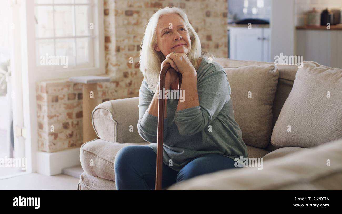 Ich Frage mich, was ich heute machen könnte. Aufnahme einer Seniorin, die zu Hause allein auf dem Sofa sitzt und nachdenklich aussieht, während sie ihren Gehstock hält. Stockfoto