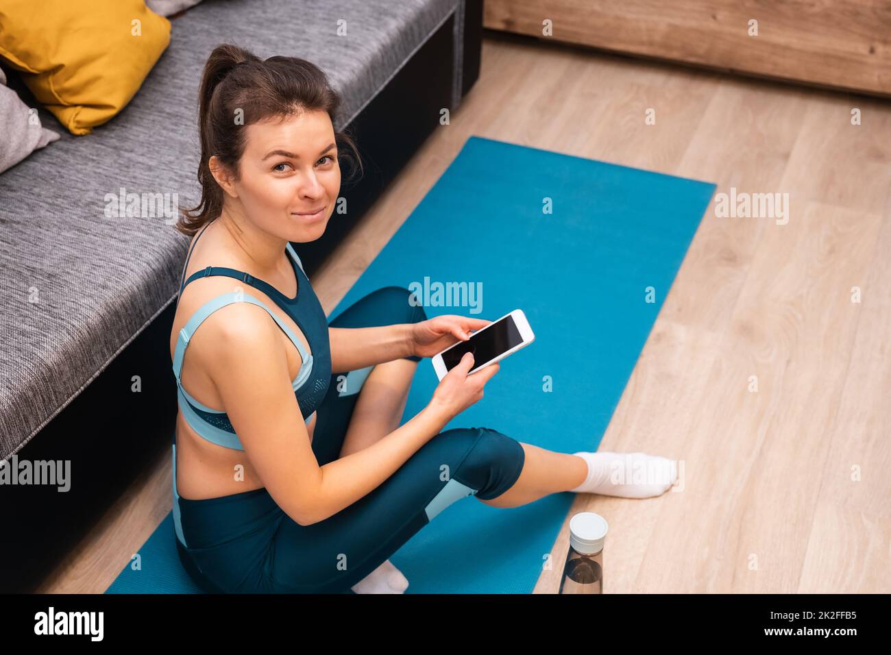 Lächelnde, athletische junge Frau mit Smartphone, während sie zu Hause auf einer Sportmatte sitzt Stockfoto