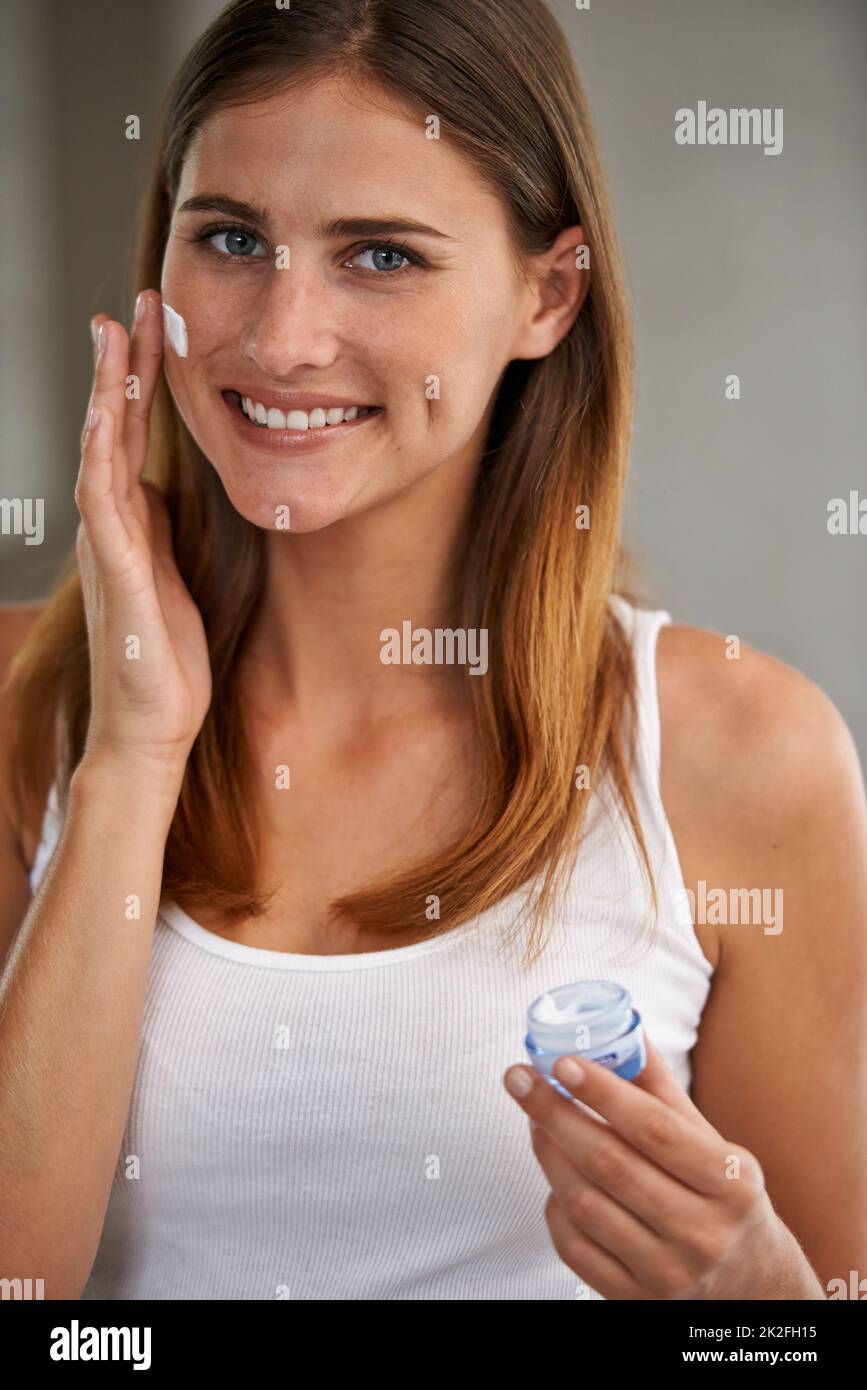 Nur ein Hauch von Feuchtigkeitscreme. Portait einer attraktiven jungen Frau, die in einem Badezimmer steht und Creme auf ihr Gesicht aufgibt. Stockfoto