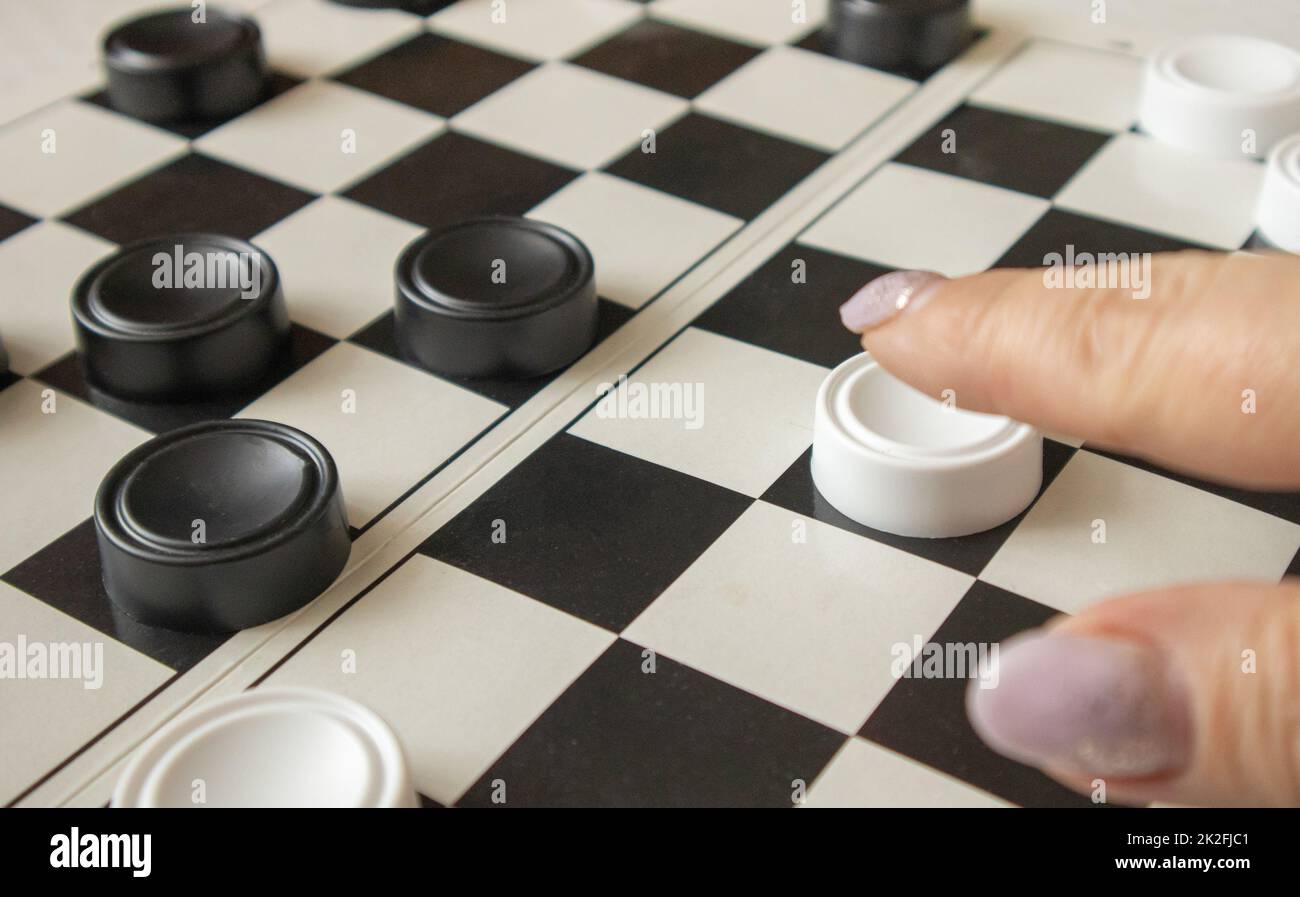 Die Hand einer Frau bewegt einen weißen Dame auf einem schwarz-weißen Spielfeld, das Konzept von Hobbys und Heimspielen Stockfoto