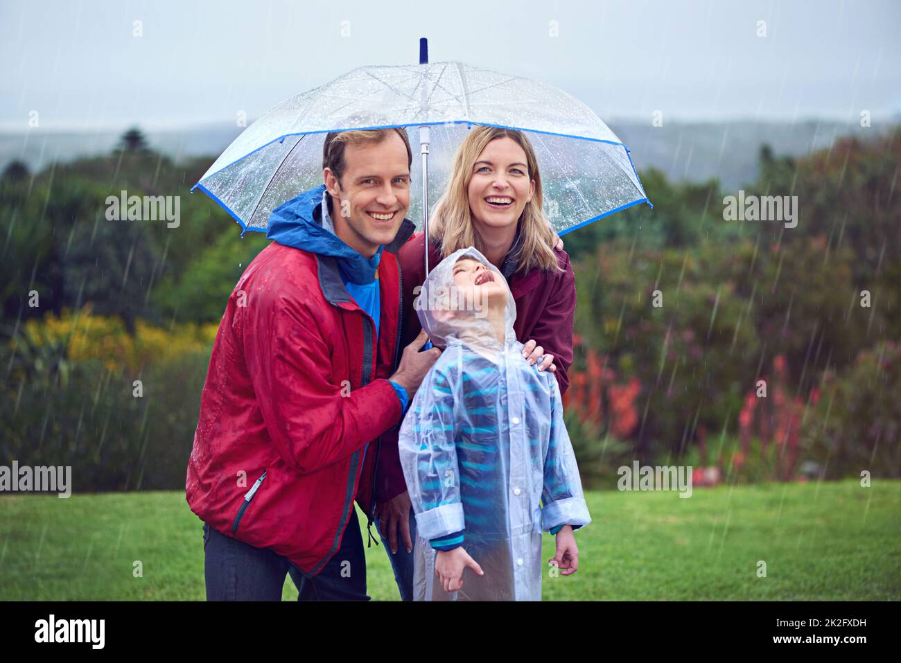 Abenteuer am Regentag. Beschnittenes Porträt einer dreiköpfigen Familie, die draußen im Regen steht. Stockfoto