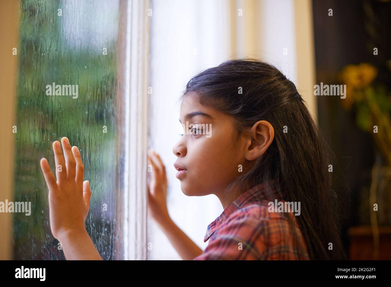 Ich hoffe, es hört auf zu regnen, damit ich nach draußen gehen und spielen kann. Aufnahme eines niedlichen kleinen Mädchens, das an einem regnerischen Tag aus dem Fenster schaut. Stockfoto