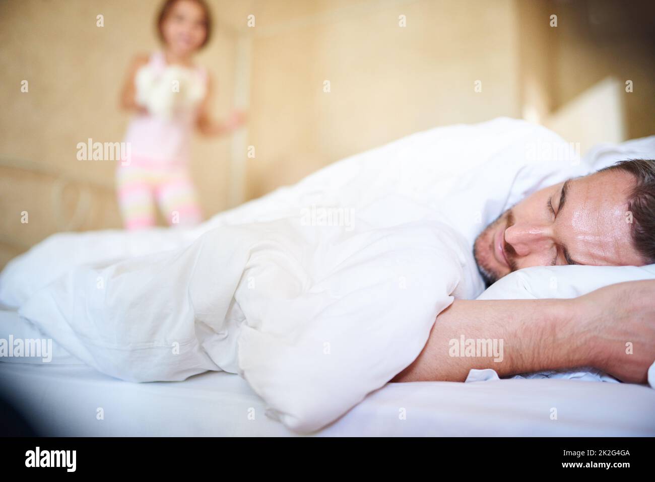 Zeit zum Aufwachen. Aufnahme eines schlafenden Vaters mit seiner kleinen Tochter, die auf das Ende des Bettes springt. Stockfoto