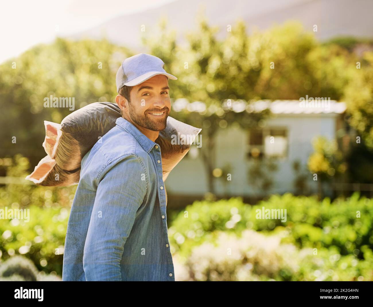 Ich habe keine Angst, meine Hände schmutzig zu machen. Porträt eines glücklichen jungen Bauern, der eine Tasche Kompost über seine Schulter auf dem Bauernhof trägt. Stockfoto