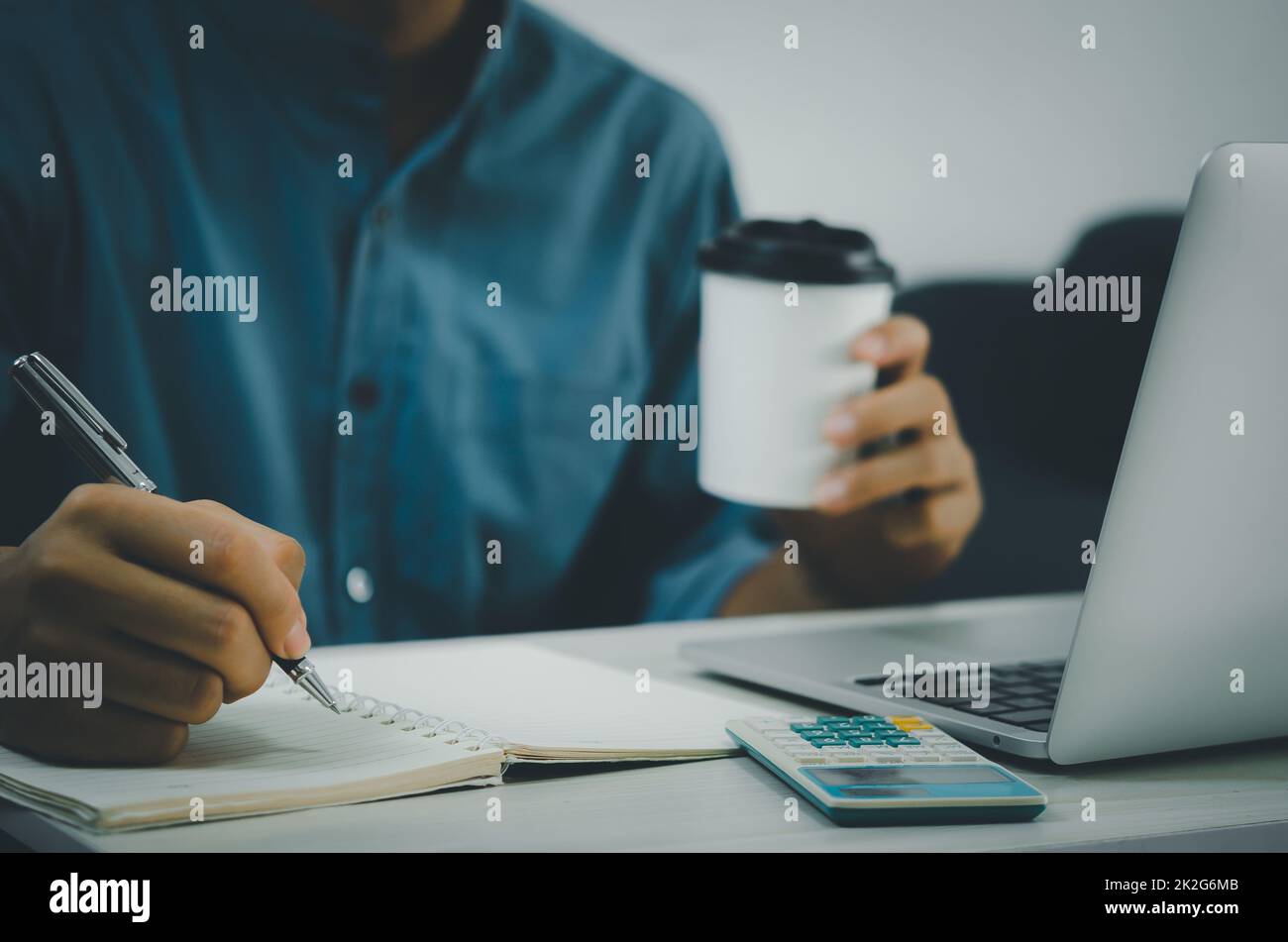 Männliche Hand hält Stift und Schreiben am Notebook und Kaffeetasse auf dem Schreibtisch mit Notebook-Computer und Rechner. Stockfoto