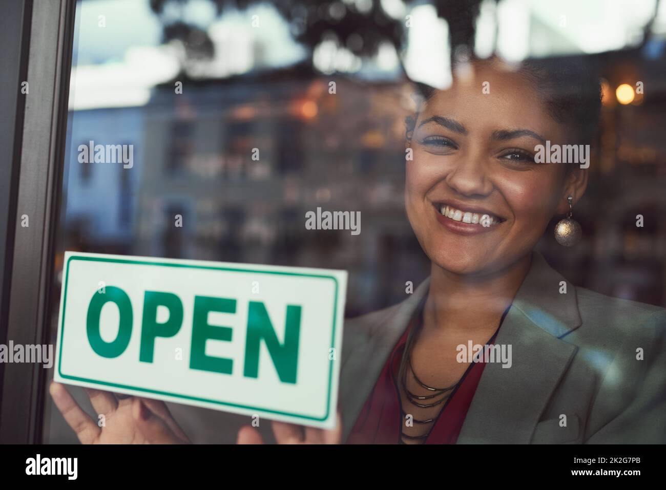 Sie freut sich darauf, alle ihre Kunden willkommen zu heißen. Porträt einer jungen Geschäftsbesitzerin, die ein offenes Schild am Fenster ihres Cafés aufgehängt hat. Stockfoto