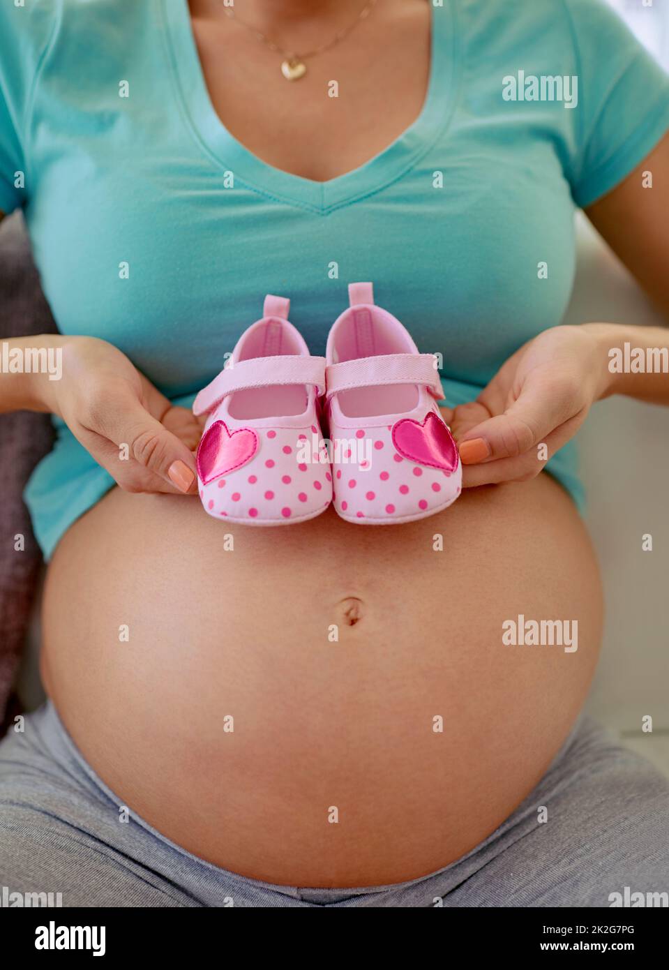 Niedliche Schuhe für meine kostbare kleine Prinzessin. Nahaufnahme einer Schwangeren, die zu Hause ein Paar rosa Babyschuhe gegen ihren Bauch hält. Stockfoto