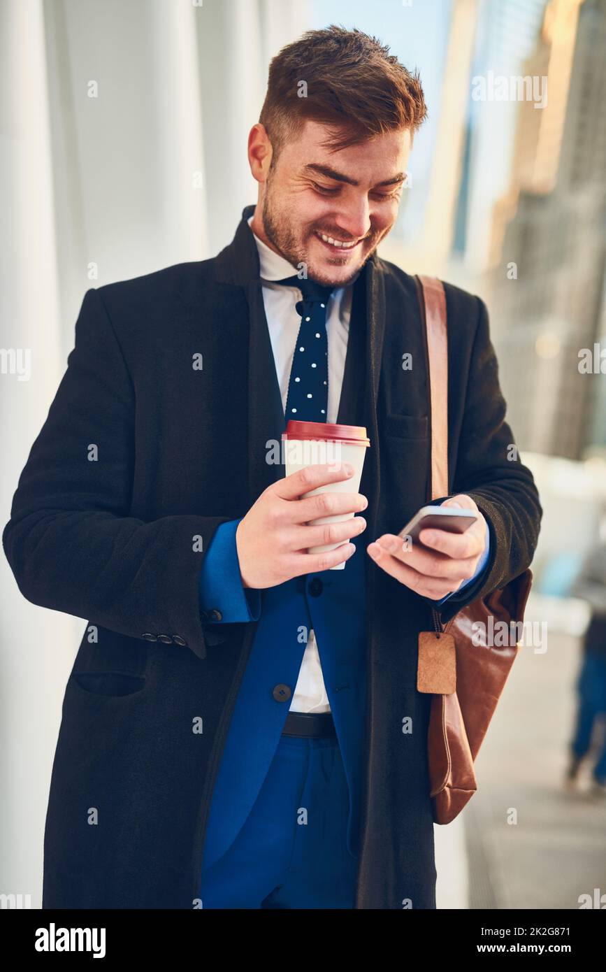 Der richtige Start in den Tag. Aufnahme eines fröhlichen jungen Mannes, der auf seinem Handy SMS schreibt, während er in den geschäftigen Straßen der Stadt Kaffee trinkt, während er morgens zur Arbeit geht. Stockfoto