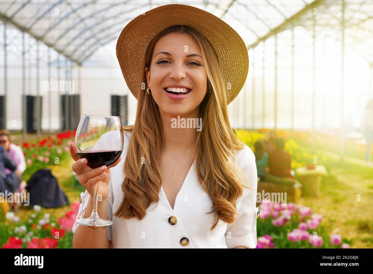 Frau, die die Kamera anschaut und ein Glas Wein mit Menschen zwischen Blumenfeldern hält, die im Hintergrund trinken und plaudern Stockfoto