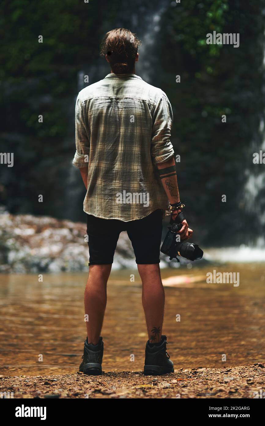 Ich möchte Fotos von allem machen. Aufnahme eines jungen Mannes, der mit umgedrehter Rückenlehne einen Wasserfall anstarrt und seine Kamera hält. Stockfoto