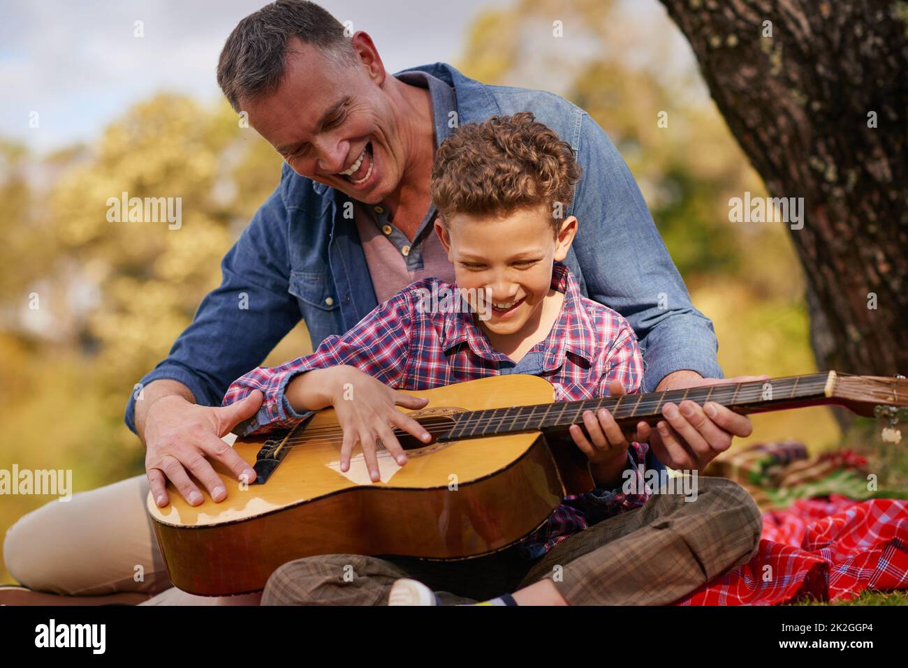 Ihm eine Melodie beibringen. Aufnahme eines Vaters, der seinem Sohn beibringt, Gitarre zu spielen, während er draußen sitzt. Stockfoto