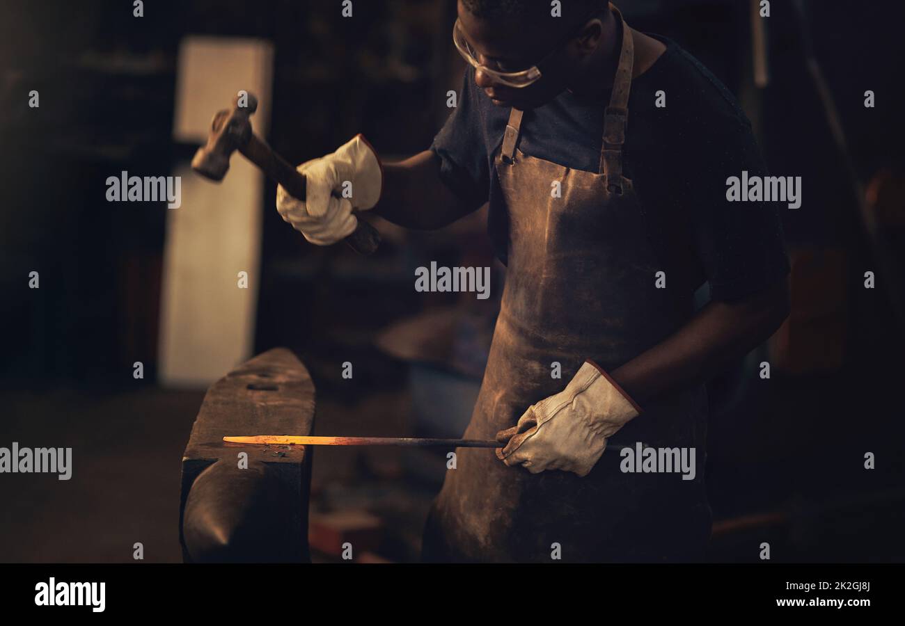 Erzielen Sie den Erfolg, den Sie verdienen. Schuss eines jungen Mannes, der mit einem Hammer in einer Gießerei auf einen heißen Metallstab schlug. Stockfoto