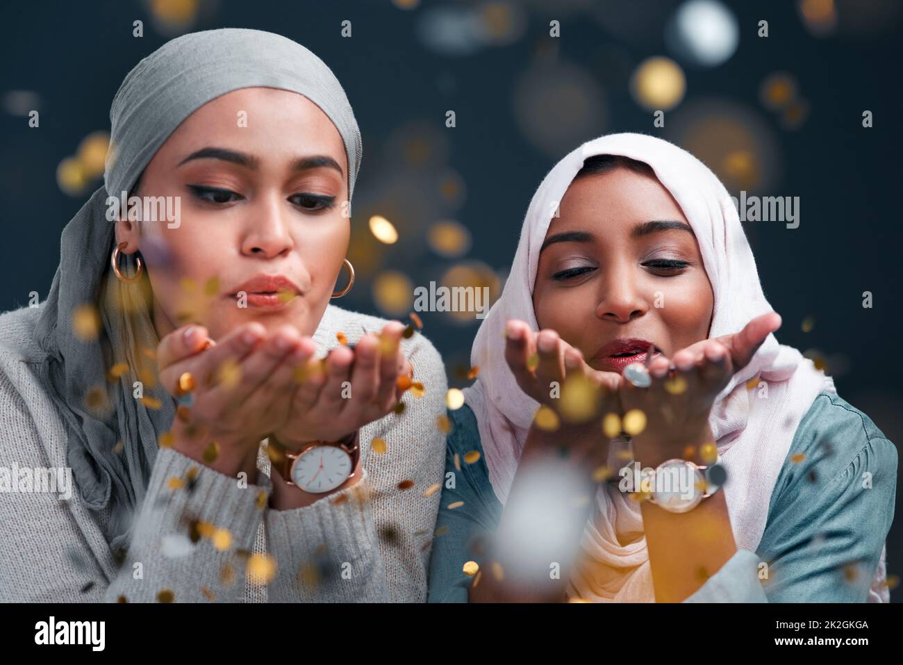 Hinterlassen Sie überall ein wenig Glitzer. Kurze Aufnahme von zwei attraktiven jungen Frauen, die Hijabs tragen und Konfetti blasen, während sie vor einem schwarzen Studiohintergrund stehen. Stockfoto