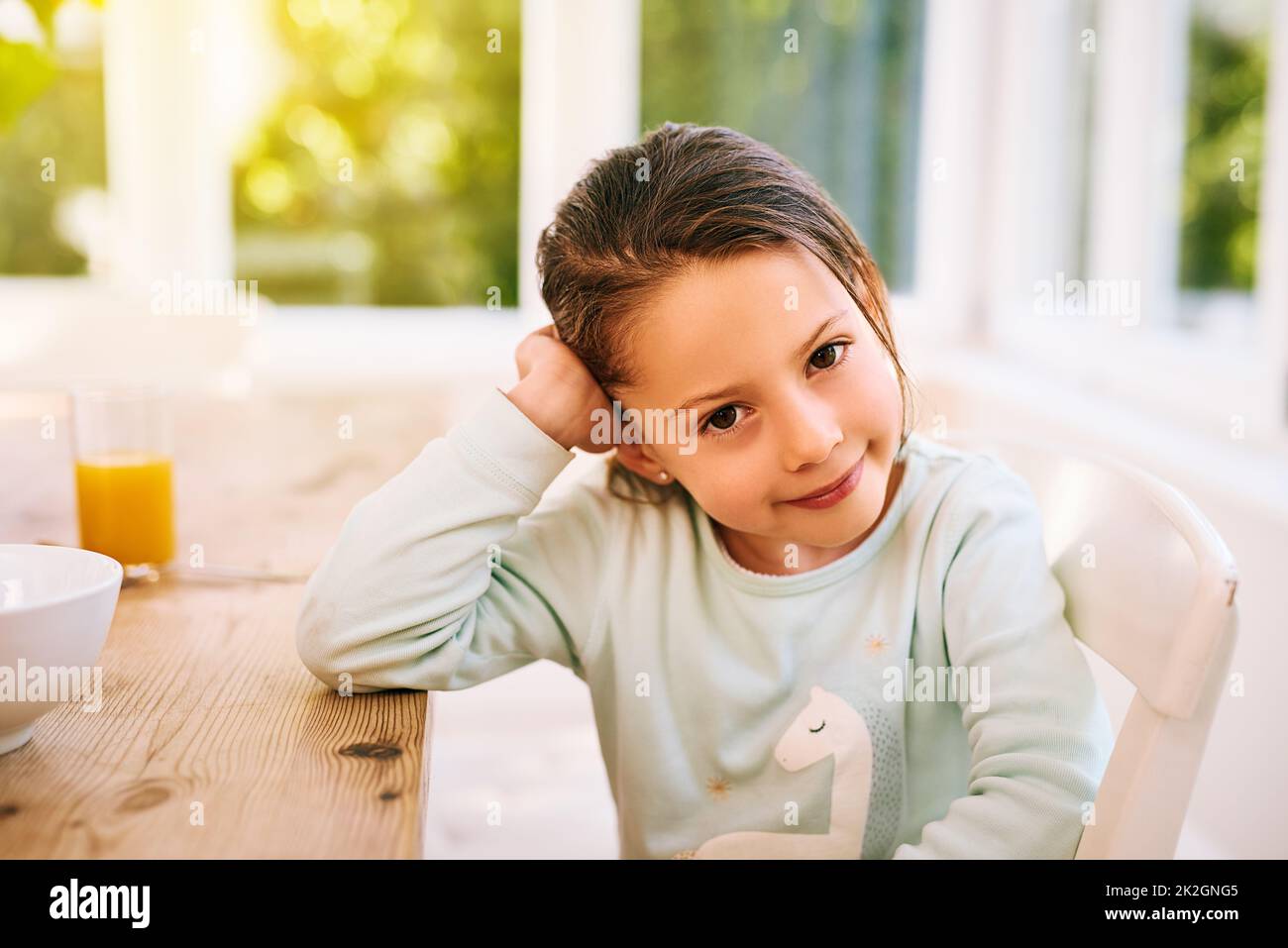 Ich will draußen spielen. Porträt eines fröhlichen kleinen Mädchens, das neben einem Küchentisch sitzt, mit einer Hand neben dem Kopf, während es in die Kamera schaut. Stockfoto