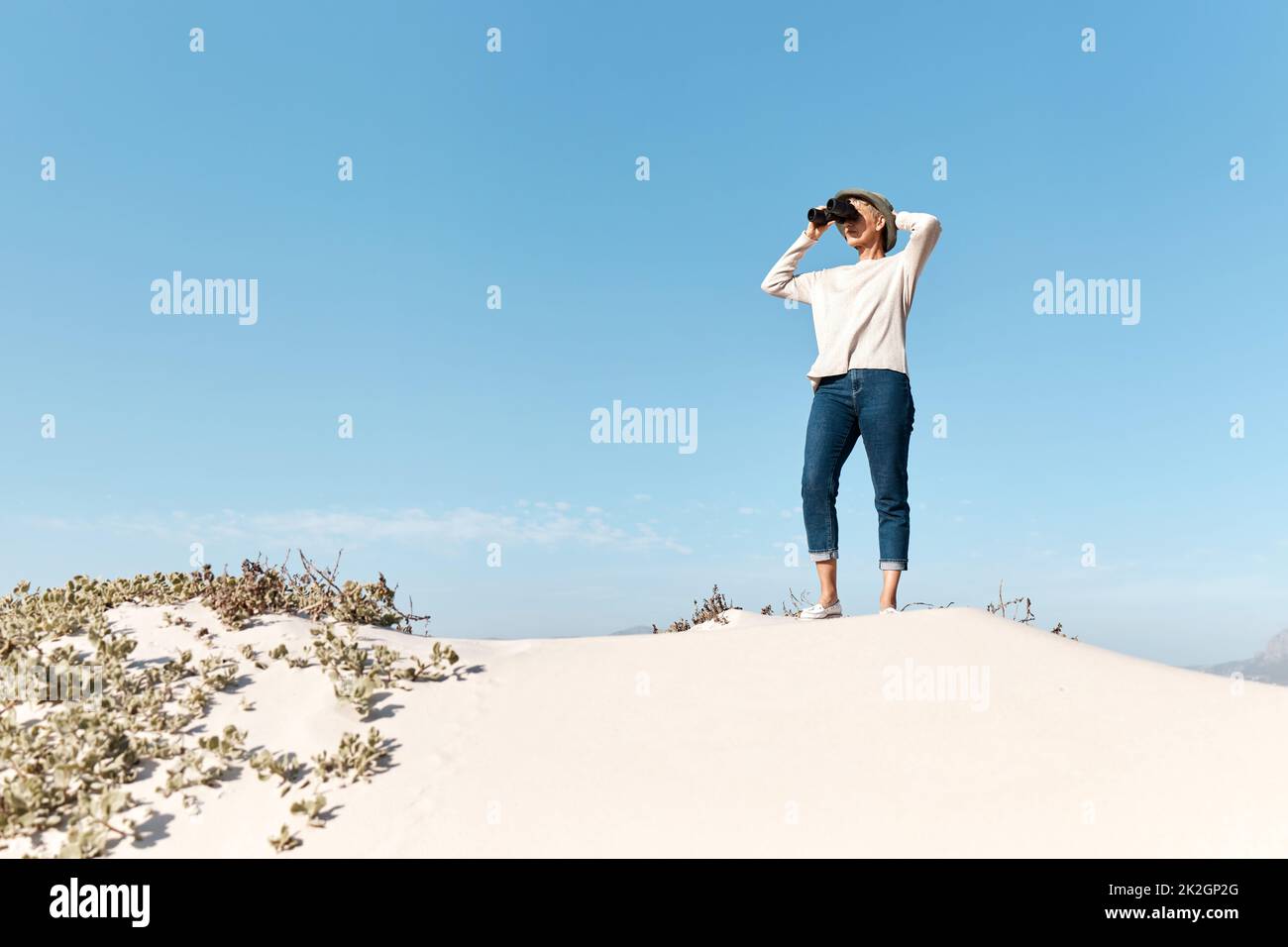 Es gibt so viel zu sehen. Ganzkörperaufnahme einer attraktiven reifen Frau, die ihr Fernglas benutzt, während sie am Strand steht. Stockfoto