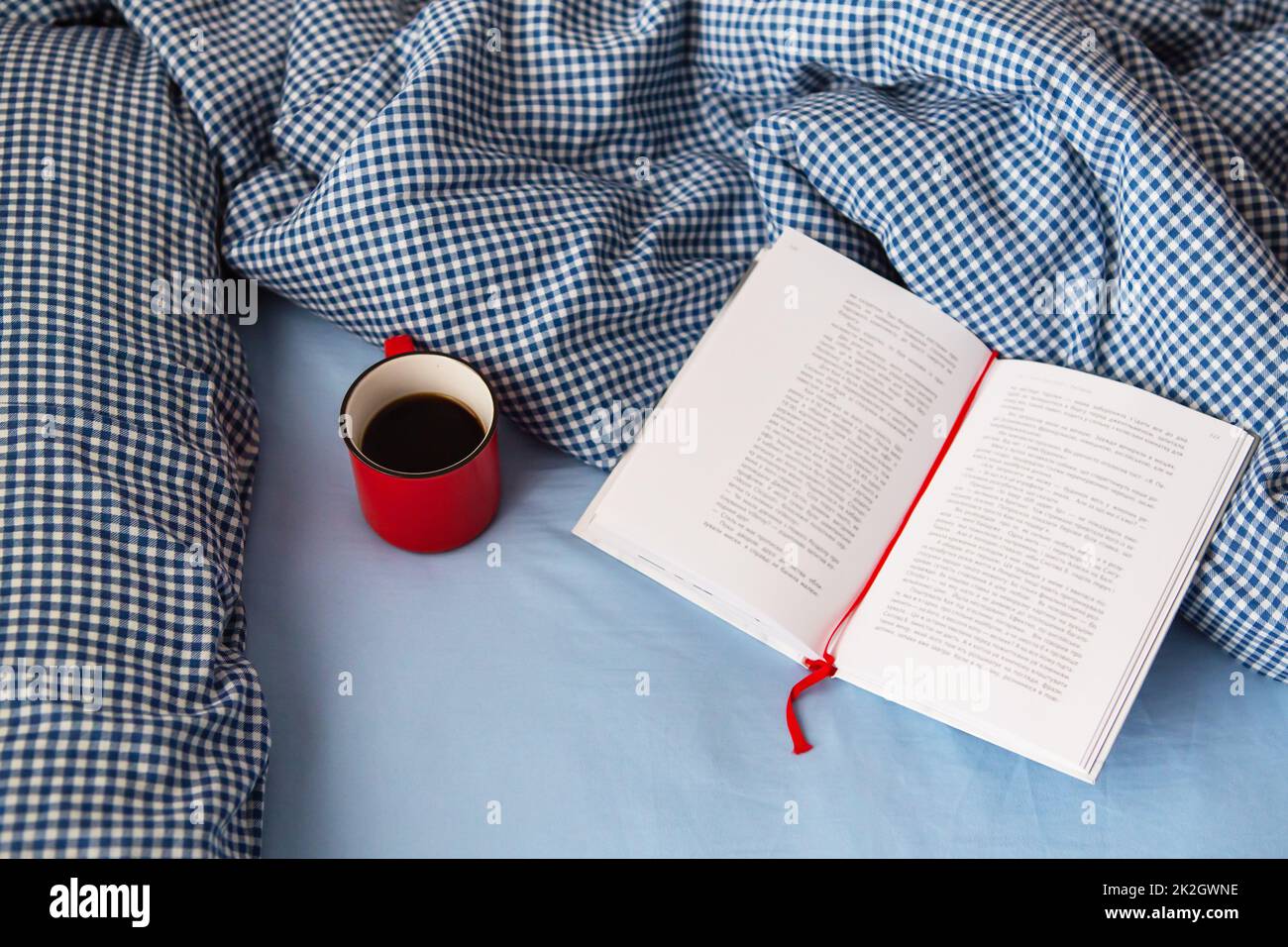 Ukraine, Rivne - 11. August 2021: Blick von oben auf eine gemütliche Decke, ein offenes Buch und eine heiße rote Tasse Kaffee auf dem Bett an einem kalten Wintertag. Entspannungs- und Hyggenkonzept. Draufsicht mit Kopierbereich. Stockfoto