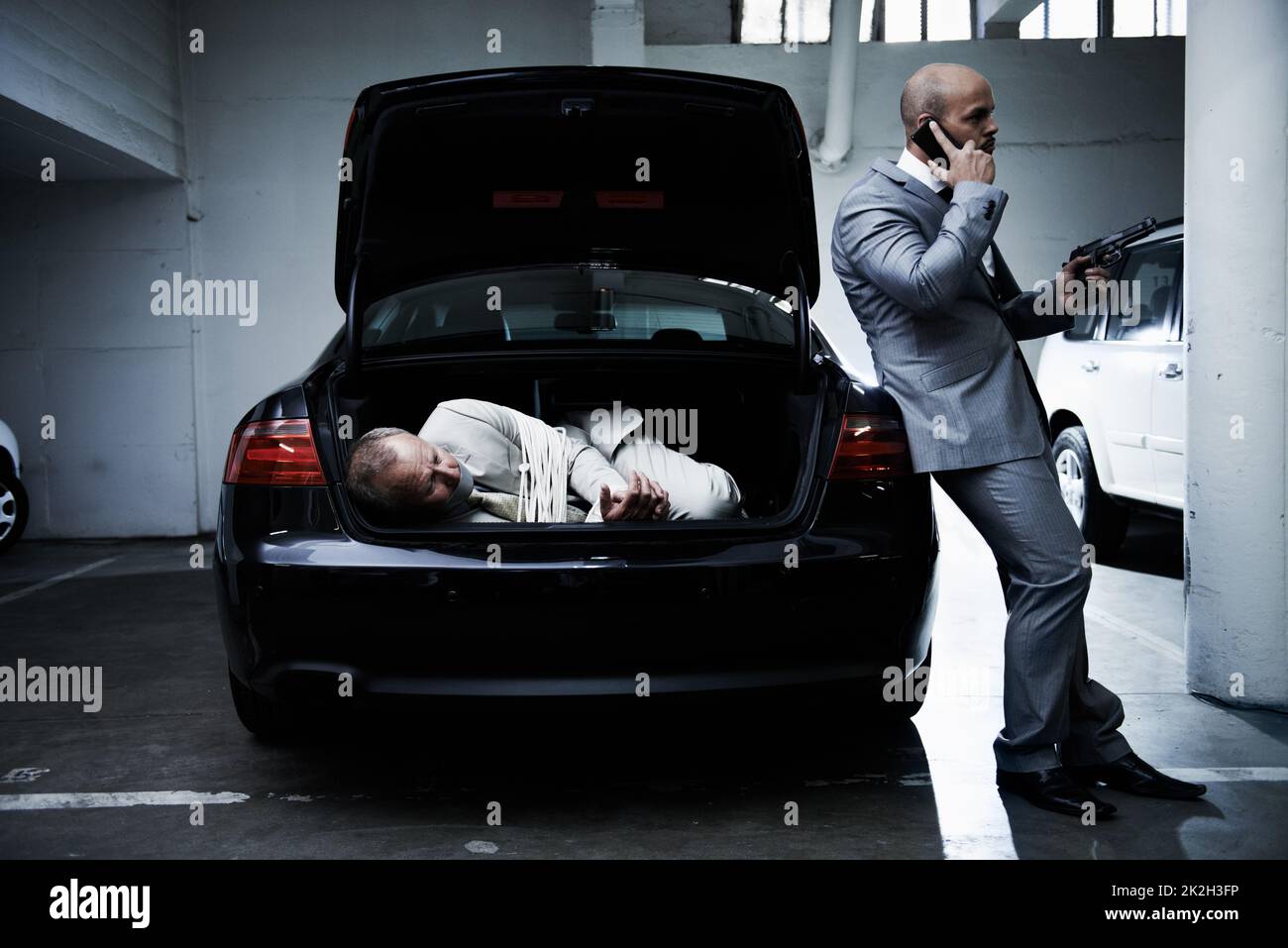 Geisel für den Mob. Ein verängstigter Geschäftsmann liegt im Kofferraum eines Autos, während sein bewaffneter Kopfgeldjäger auf einem Handy spricht. Stockfoto