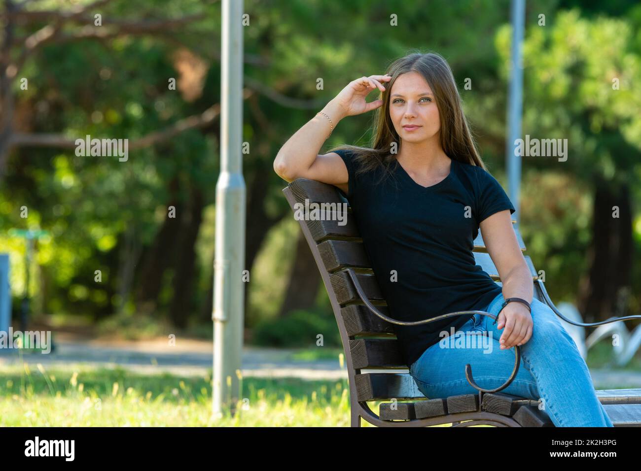 Ein schönes Mädchen in legerer Kleidung sitzt auf einer Bank in einem schönen grünen Park, das Mädchen schaut in den Rahmen Stockfoto