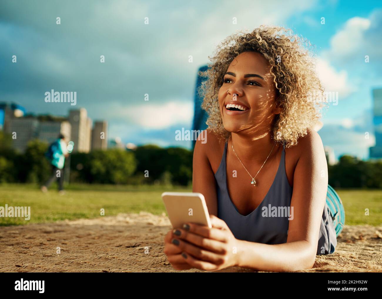 Die Sonne, der Park und das perfekte E-Book. Ausgeschnittene Aufnahme einer attraktiven jungen Frau, die ein Smartphone benutzt, während sie sich in einem Park niederlegt. Stockfoto