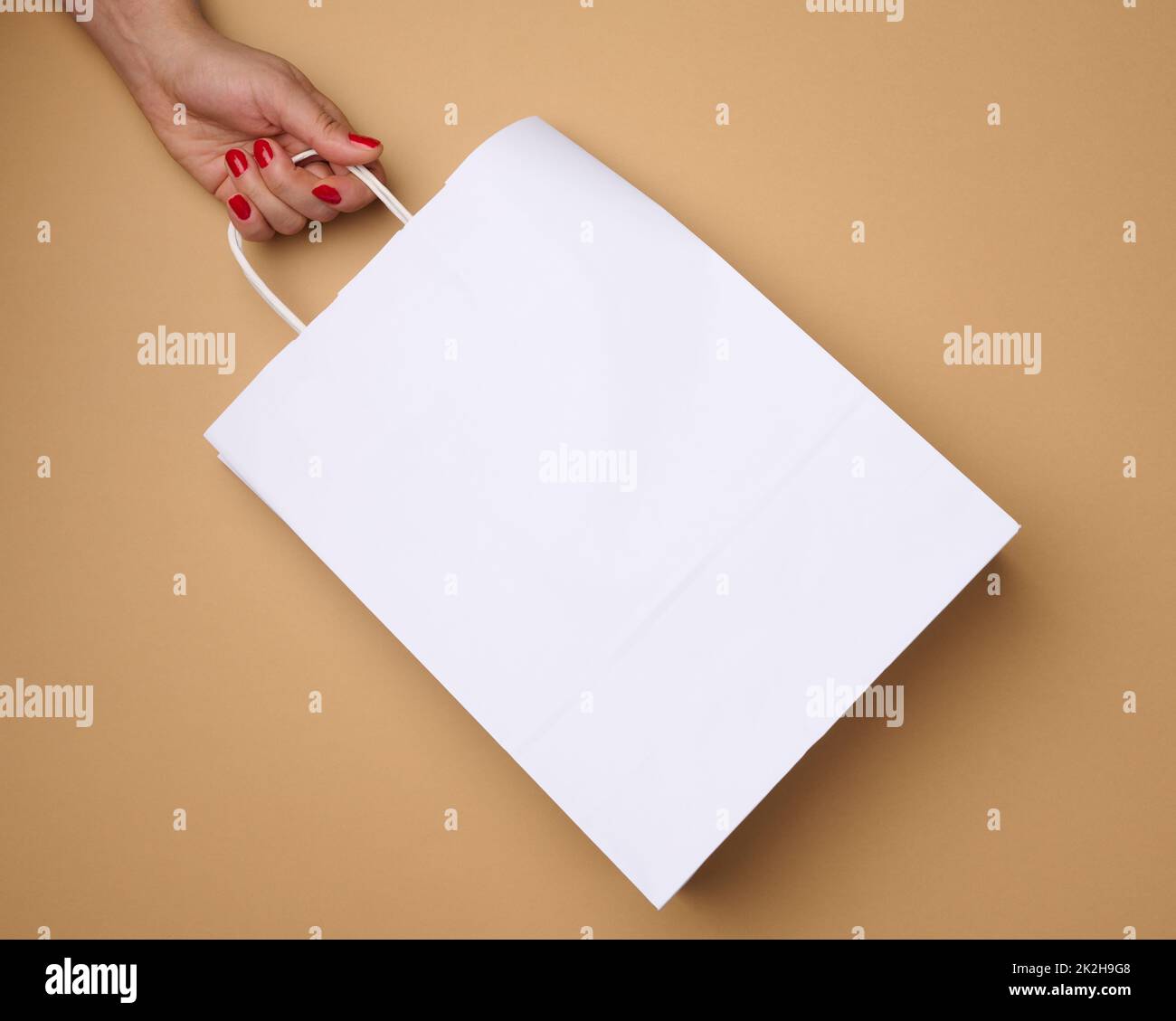 Weibliche Hand hält eine leere weiße Papiertüte an den Griffen auf braunem Hintergrund, Ablehnung von Plastiktüten Stockfoto