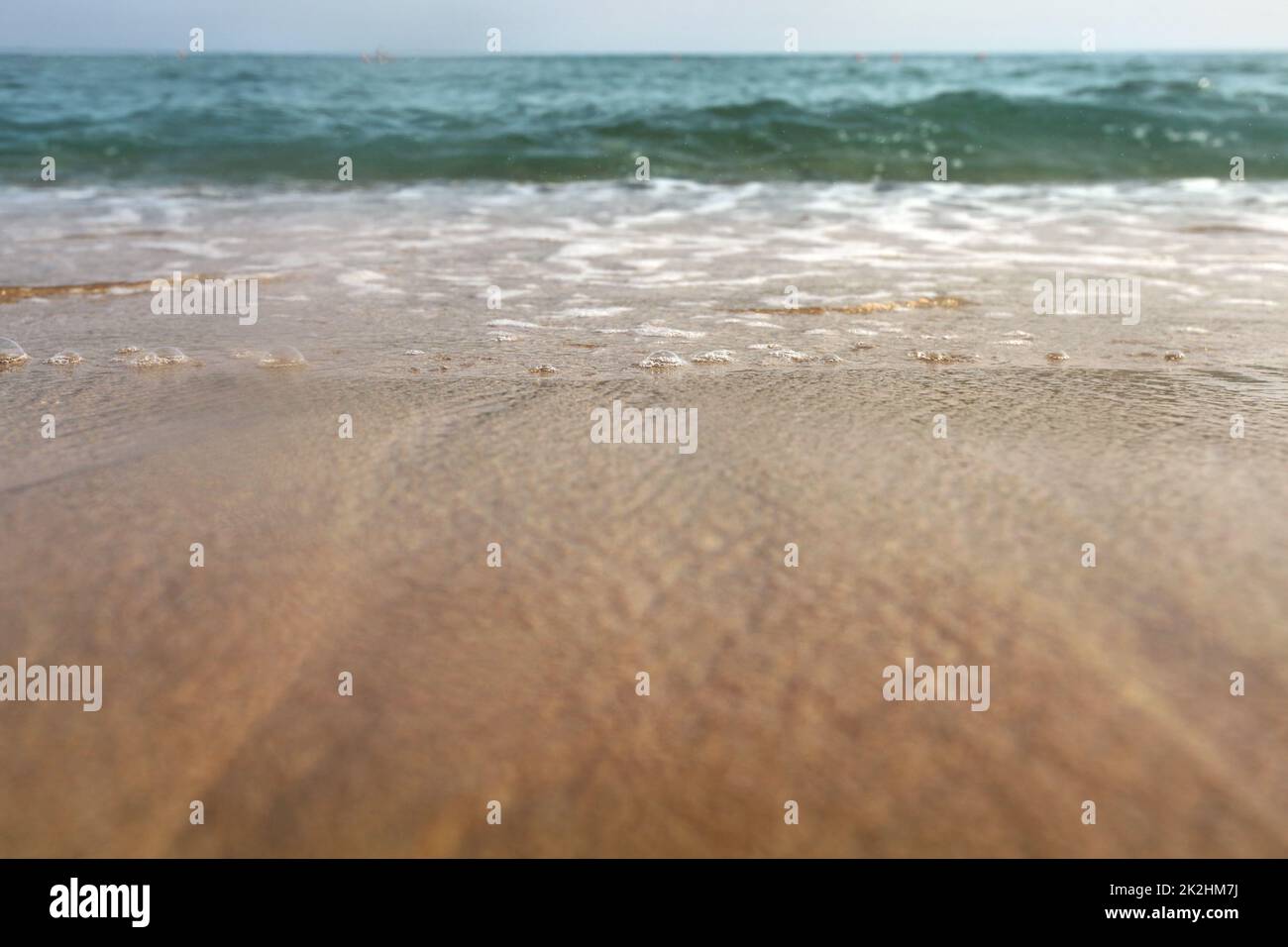 Flachwinkelfoto vom Erdgeschoss - Strand Sand nass vom Meer, Wassertropfen in der Luft, kleine Wellen und verschwommenes Meer in der Ferne. Abstrakter Urlaubshintergrund. Stockfoto
