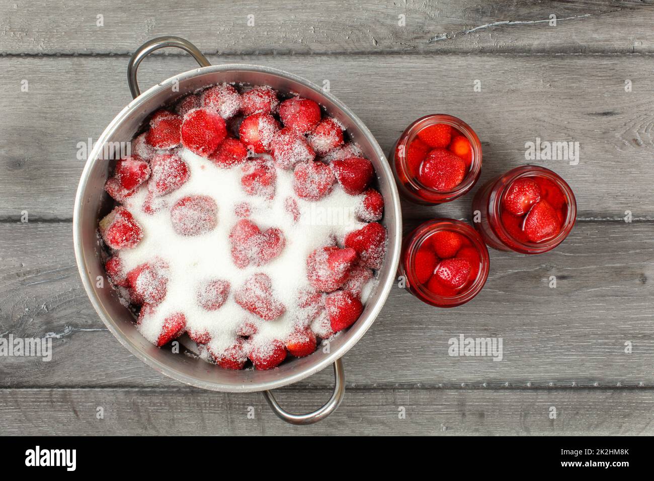 Blick auf den Tisch - Stahl mit Erdbeeren bedeckt mit Kristallzucker, drei Flaschen mit eingelegter Erdbeere daneben. Zubereitung von selbstgemachter Marmelade/Kompott Stockfoto