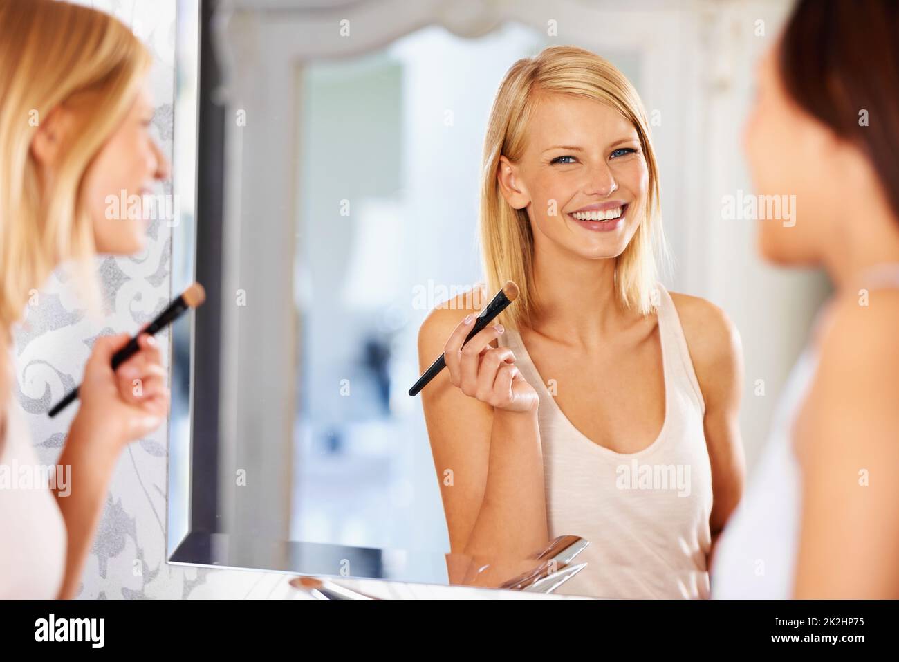 Ihre Haut sieht strahlend schön aus. Eine junge Frau, die sich vor einem Spiegel schminkt, während ihre Freundin dabei steht. Stockfoto