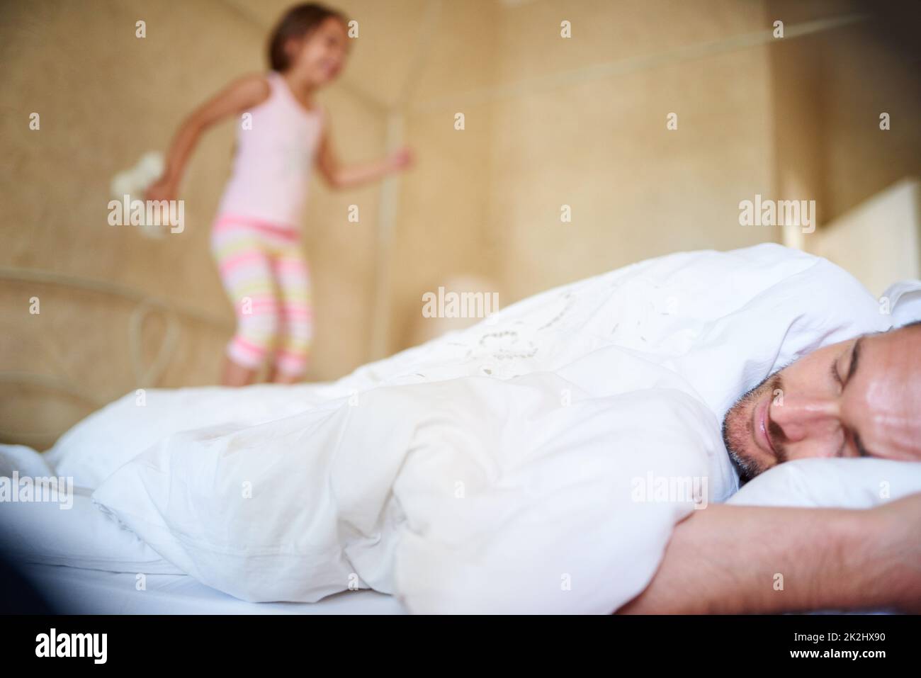 Zeit zum Aufwachen. Aufnahme eines schlafenden Vaters mit seiner kleinen Tochter, die auf das Ende des Bettes springt. Stockfoto