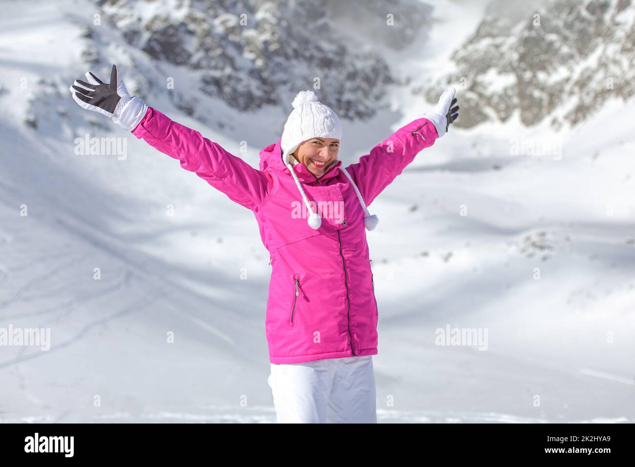 Junge Frau in rosa Winter Ski Jacke, Mütze und Handschuhe, die Arme ausgebreitet, lächelnd, glücklich mit Schnee bedeckten Berg hinter ihr. Stockfoto