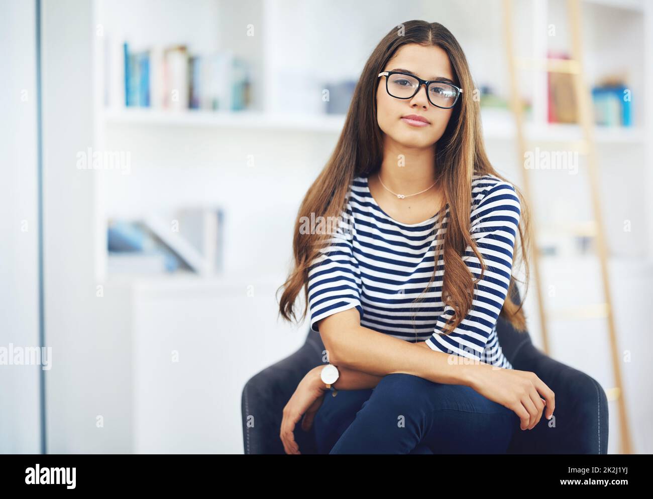 Shes voller Selbstvertrauen. Porträt einer lächelnden jungen Frau mit Brille, die zu Hause vor Bücherregalen sitzt. Stockfoto