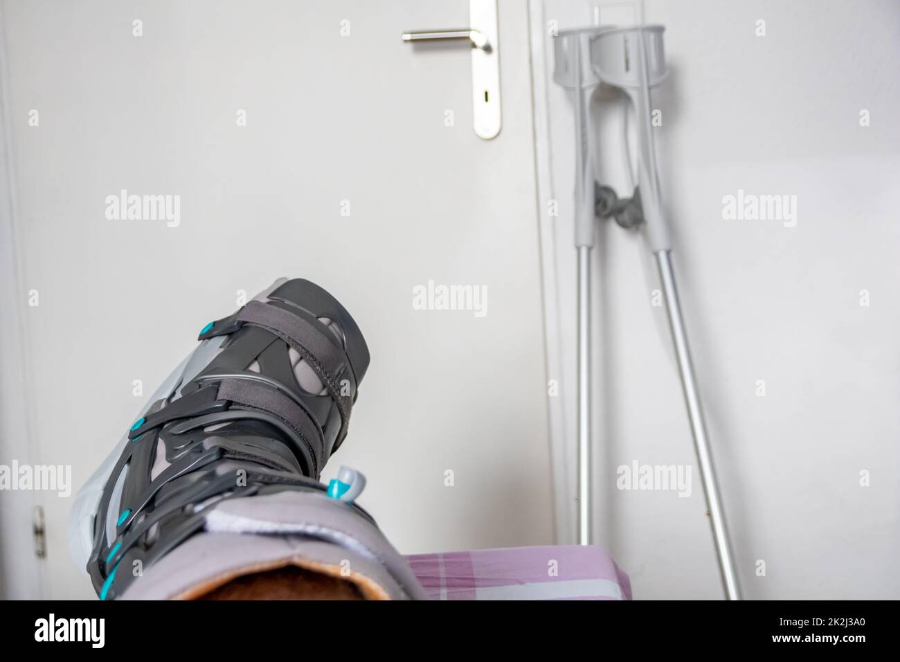 Der europäische Mann nach der Achillessehnenruptur ist wieder zu Hause mit einem speziellen Physiotherapieschuh und Krücken für die Heilung zu Hause mit gesunden Medikamenten Schmerzmittel gegen das schmerzende Bein Stockfoto