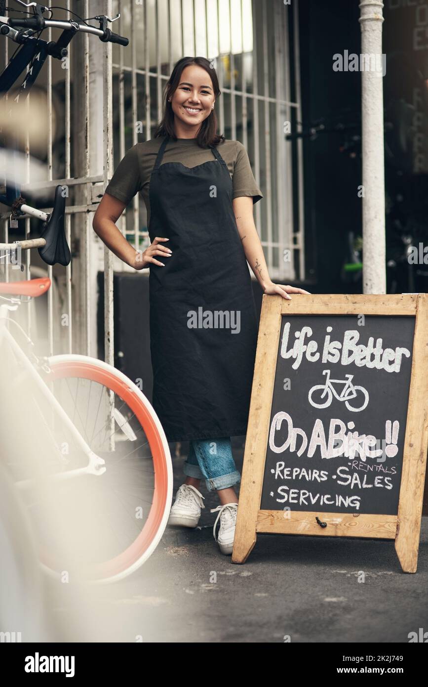 Kommen Sie herein. Ganzkörperaufnahme einer attraktiven jungen Frau, die tagsüber vor ihrem Fahrradladen steht. Stockfoto