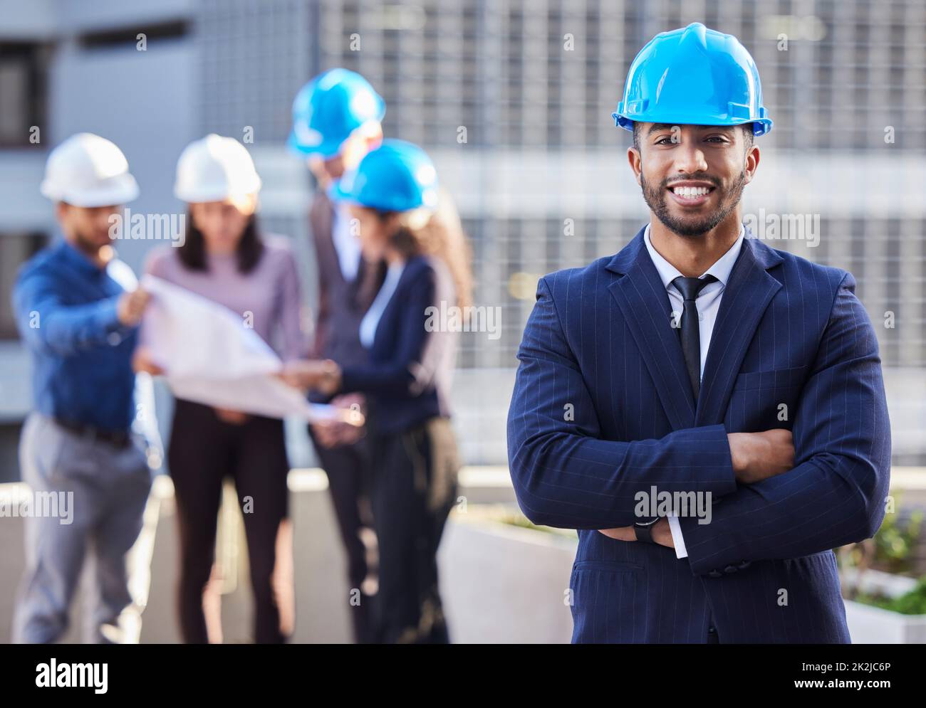 Ich habe das Team, das es möglich machen kann. Foto eines jungen Geschäftsmannes, der mit zusammengefalteten Armen steht und einen Schutzhelm trägt, während seine Kollegen hinter ihm stehen. Stockfoto