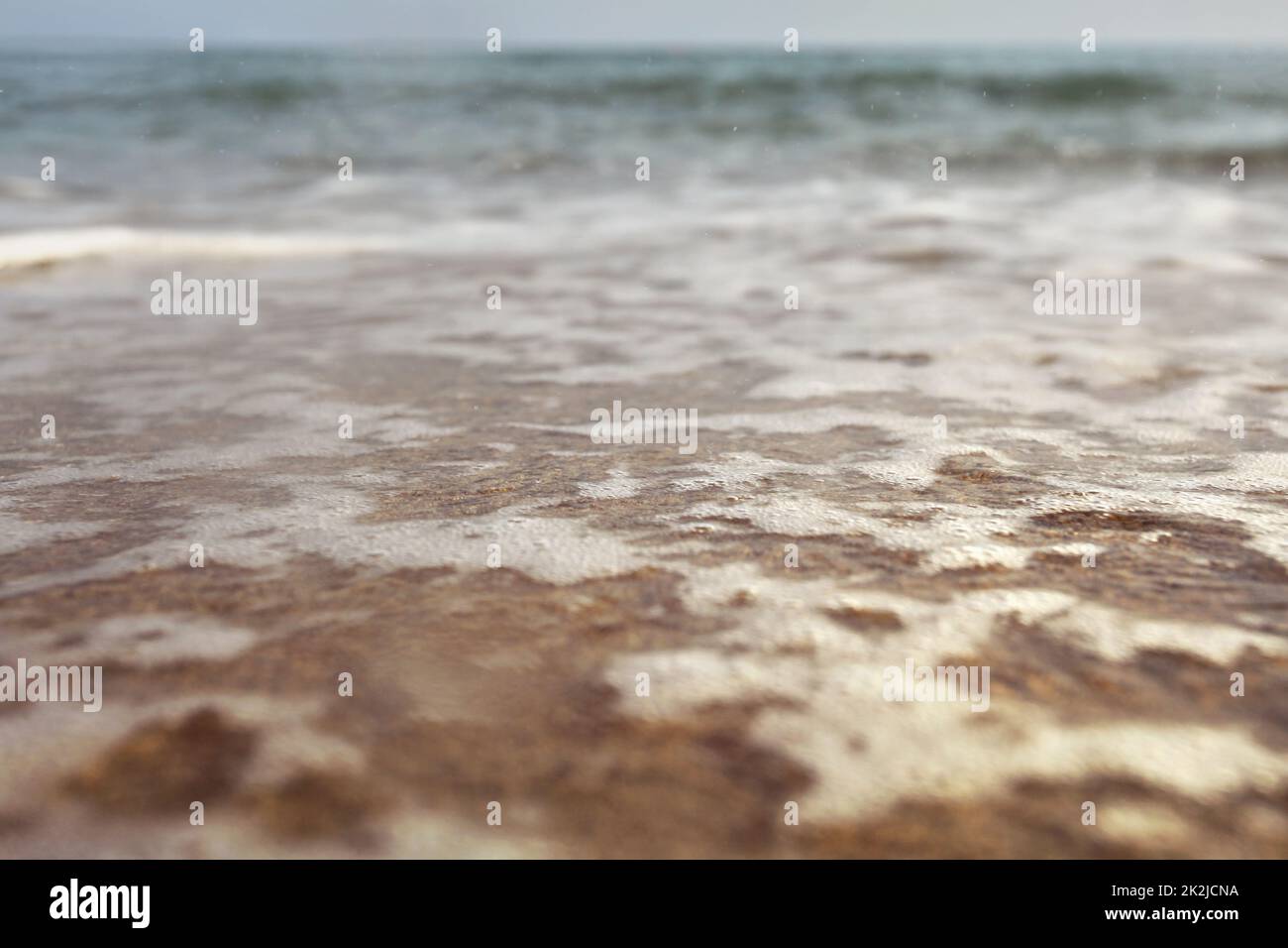 Flachwinkelfoto vom Erdgeschoss - Strand mit flachem Wasser und weißem Schaum über Sand, nasse Lufttropfen, verschwommenes Meer in der Ferne. Abstrakter Urlaubshintergrund. Stockfoto