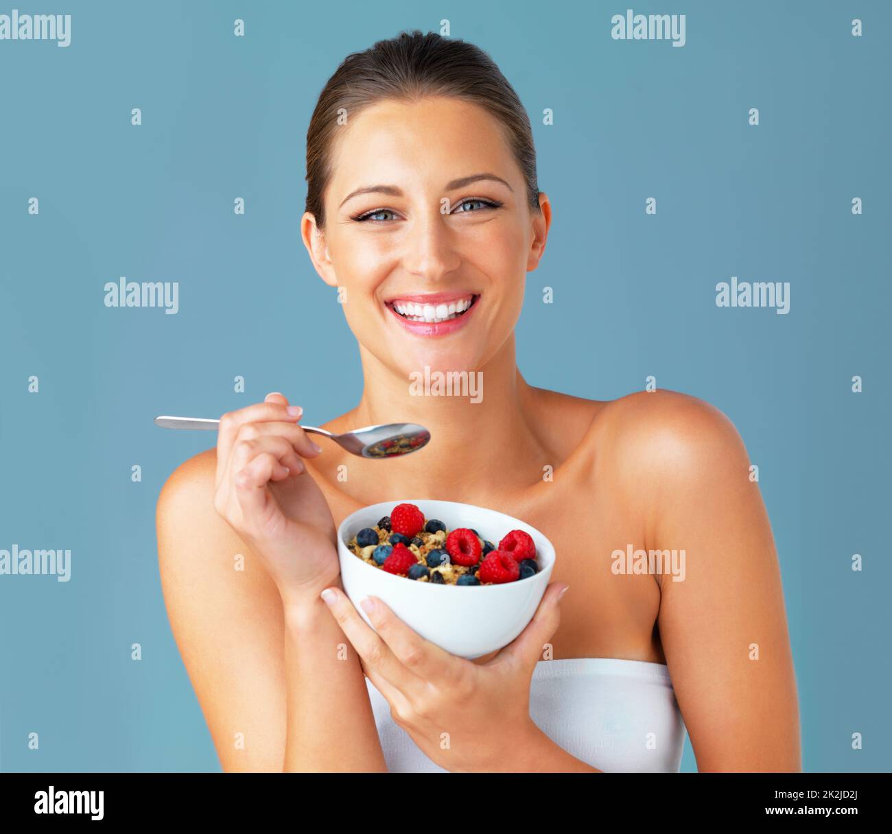 Gesundheit und Glück gehen Hand in Hand. Studioaufnahme einer attraktiven jungen Frau, die vor blauem Hintergrund eine Schale Müsli und Obst isst. Stockfoto