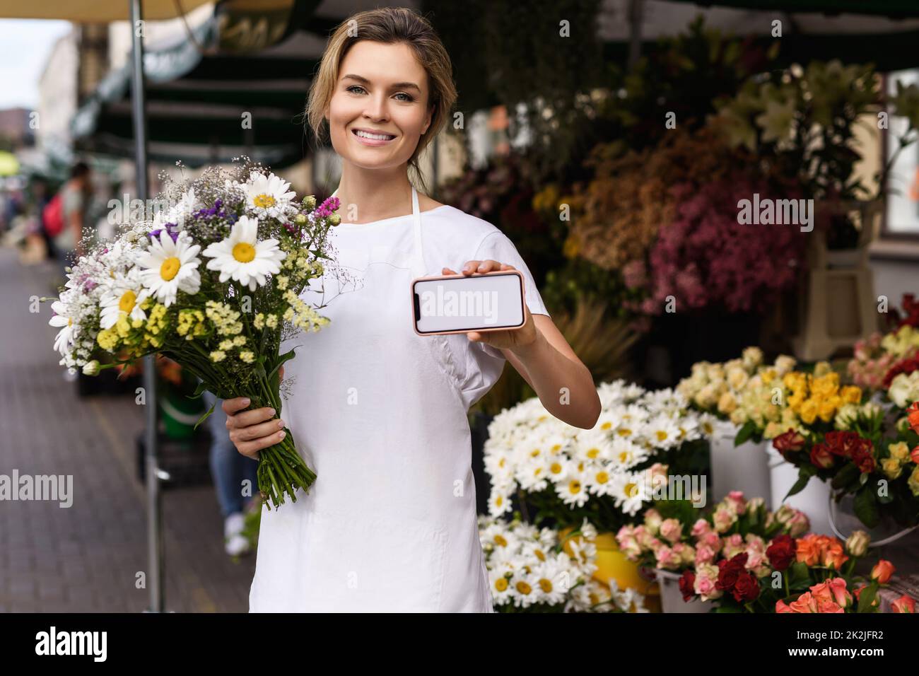 Eine Blumenfrau mit einem Blumenstrauß und einem Smartphone mit einem leeren Display in ihrem kleinen Blumenladen Stockfoto